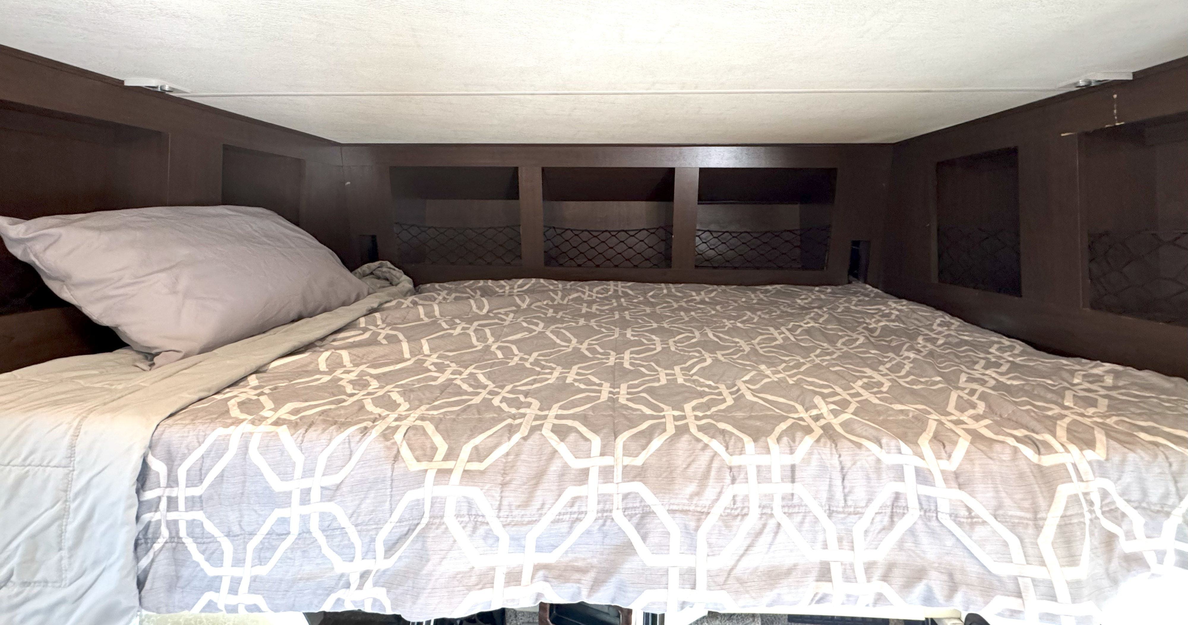 Cozy RV rental bed with a grey patterned comforter and pillow, featuring dark wood overhead storage with mesh pockets.. Thor Motor Coach A.C.E 2019