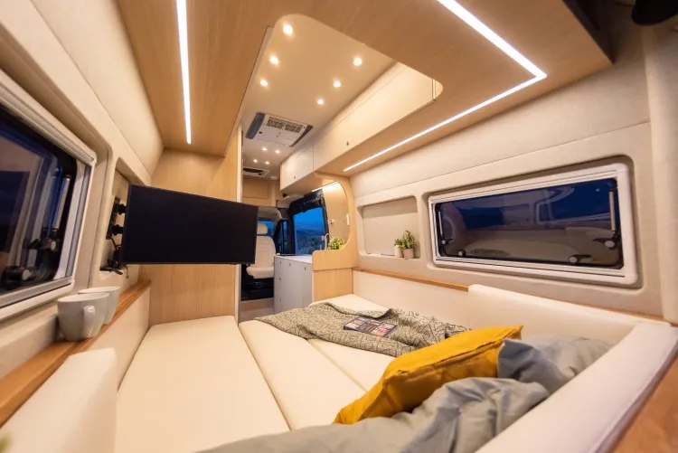 Bright, modern RV rental interior featuring a large bed/lounge, wall-mounted TV, and ample lighting. Ideal for travel.. Noovo Plus RL 2025
