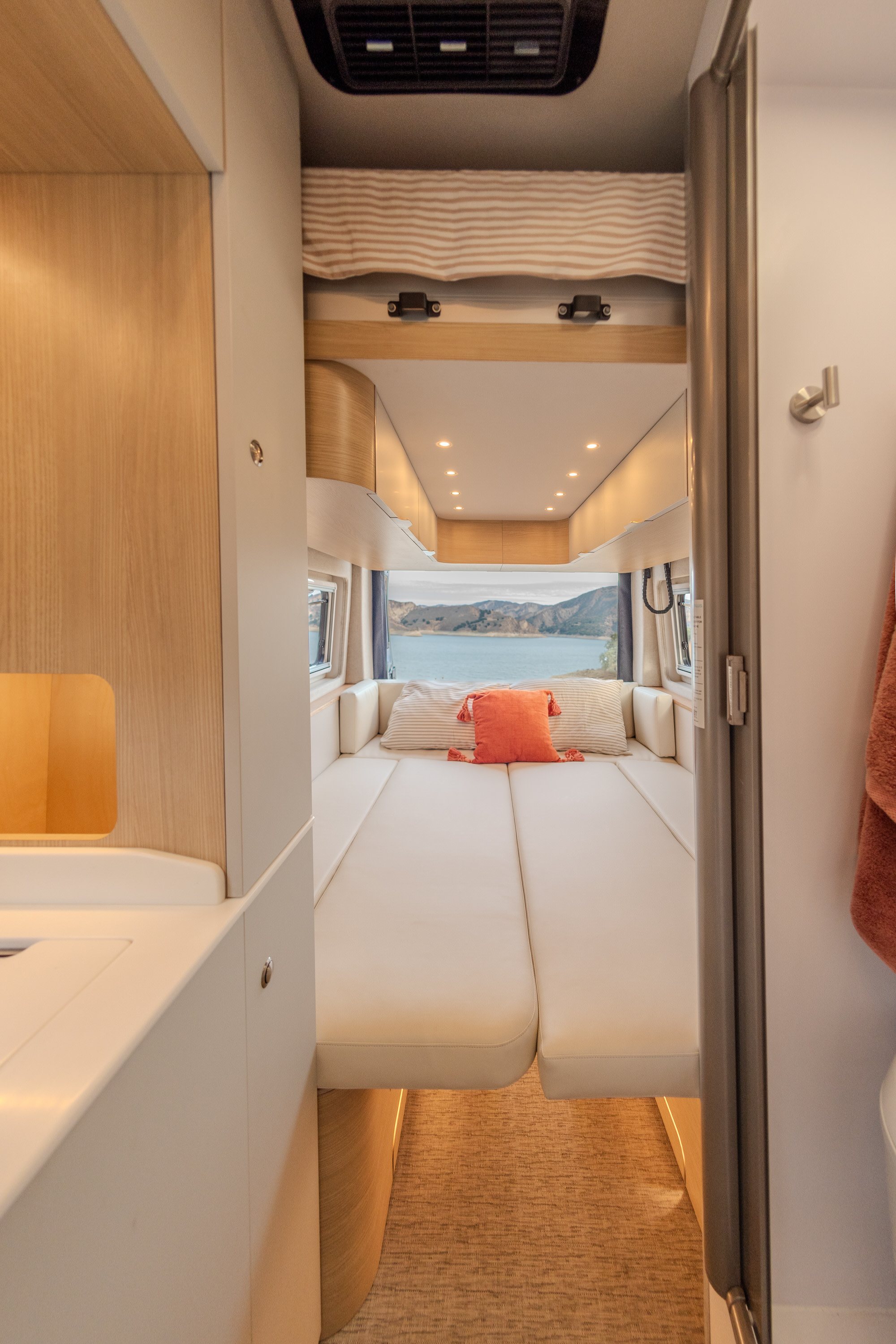 This RV rental offers a well-lit interior with a large bed and a picturesque view of a lake and mountains.. Noovo Plus 2025
