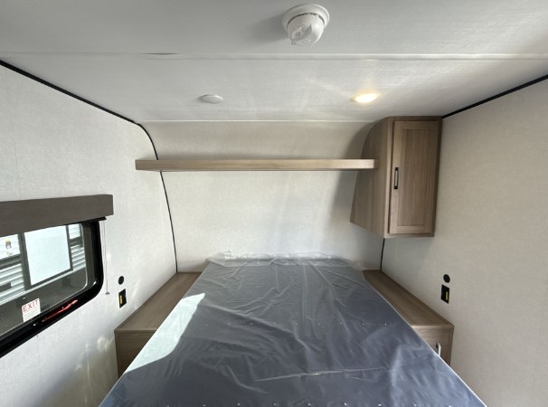 Clean RV rental bedroom features a bed with plastic covering, overhead shelf, and cabinet. Window & smoke detector visible.. Keystone Coleman 17R 2025