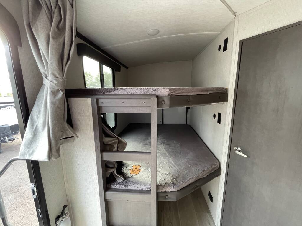Inside an RV rental: view of two grey-padded bunk beds, ideal for extra sleeping space. Curtains and a side door visible.. Keystone RV Cougar 2023