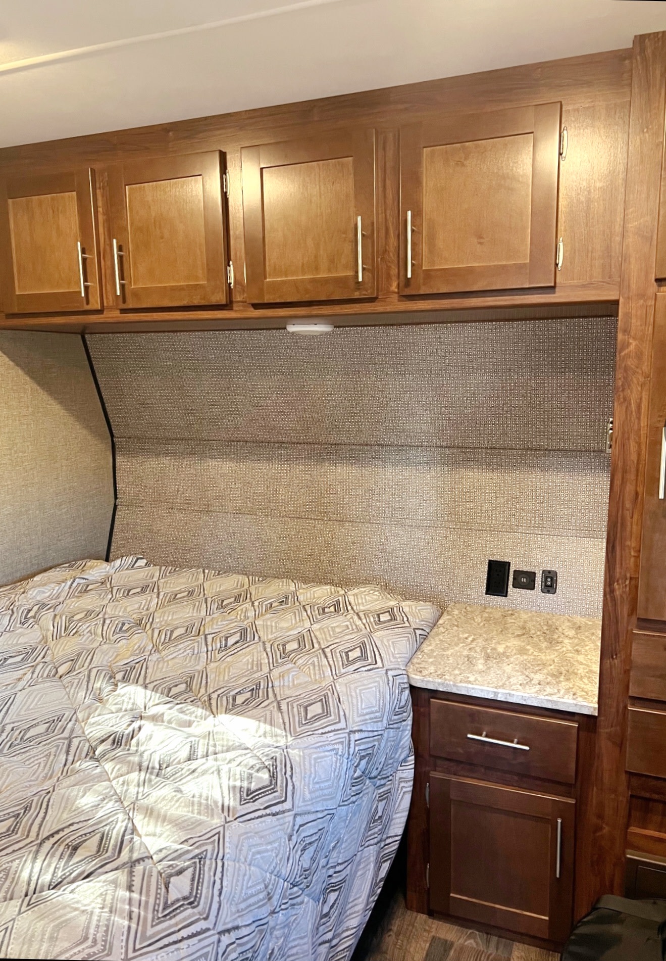Inside a cozy RV rental: wooden overhead cabinets, a patterned bed, and a compact nightstand with outlets.. Heartland Terry Classic V21 2018