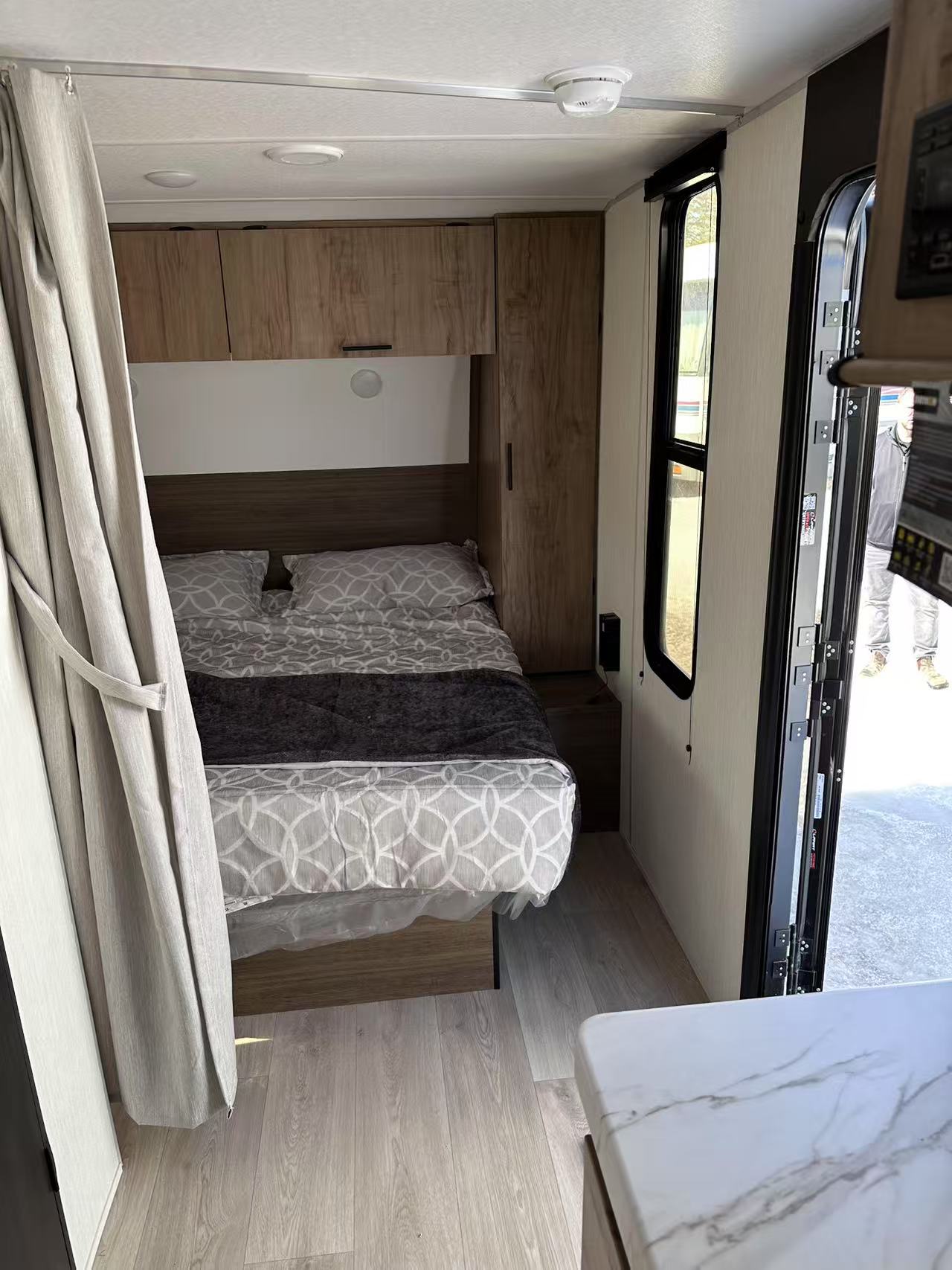 Inside an RV rental: a compact bedroom with a bed, overhead storage, light wood floor, and an open side door.. Forest River Cherokee Grey Wolf Special Edition 2025