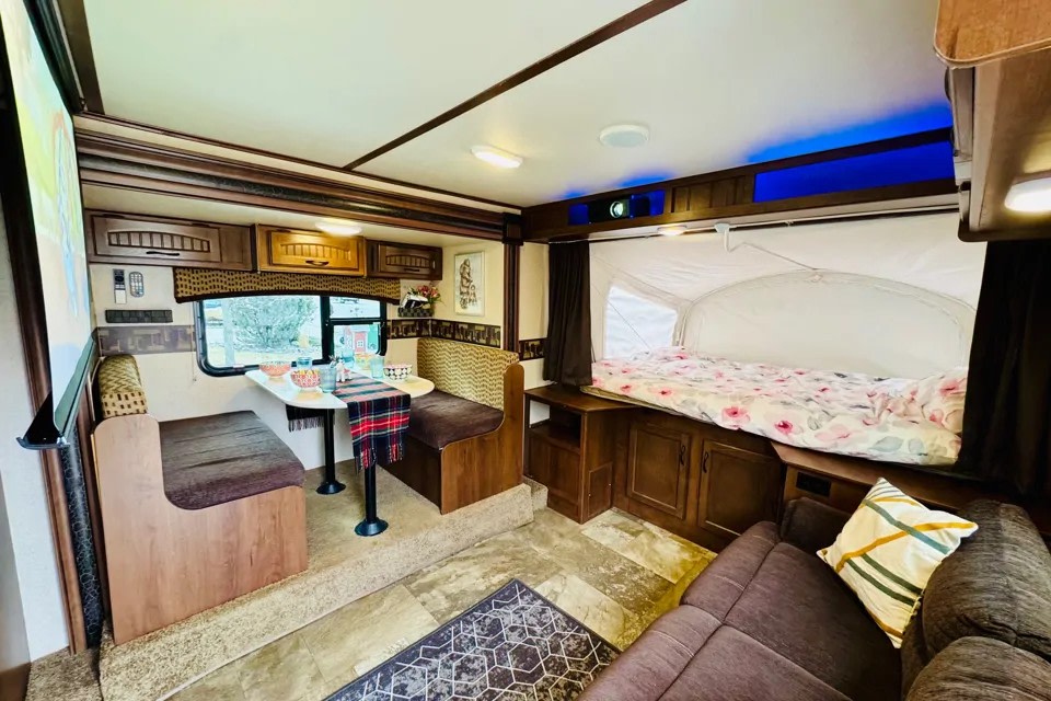 RV rental interior: Dinette, projector screen, sofa, and comfortable pop-out bed with floral comforter. Ideal for a family getaway.. Jayco Jay Feather 2018