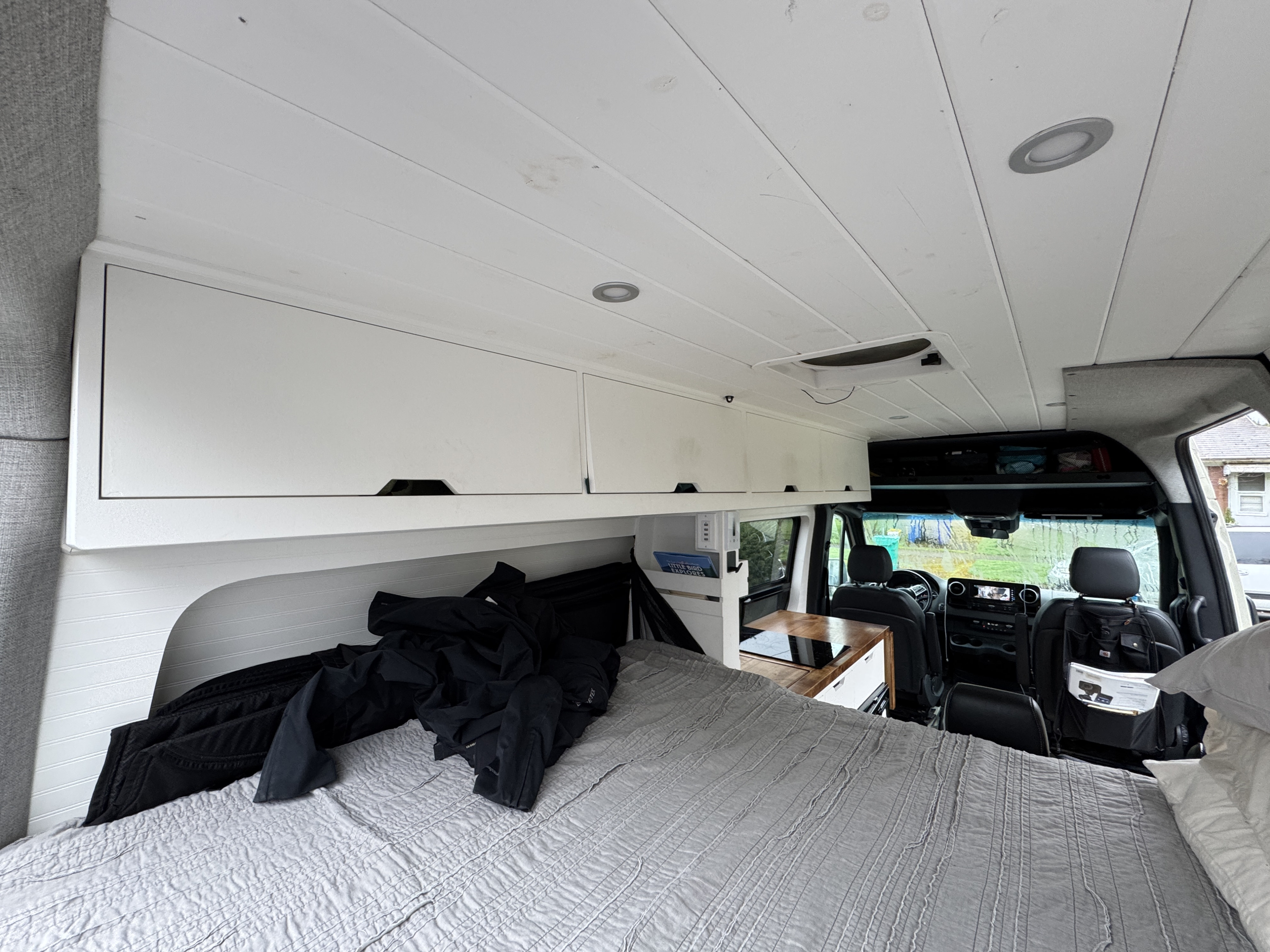 View from the back of an RV rental, showing a grey bed, white overhead cabinets, and a compact kitchen counter, leading to the front cab.. Mercedes-Benz Sprinter 2020