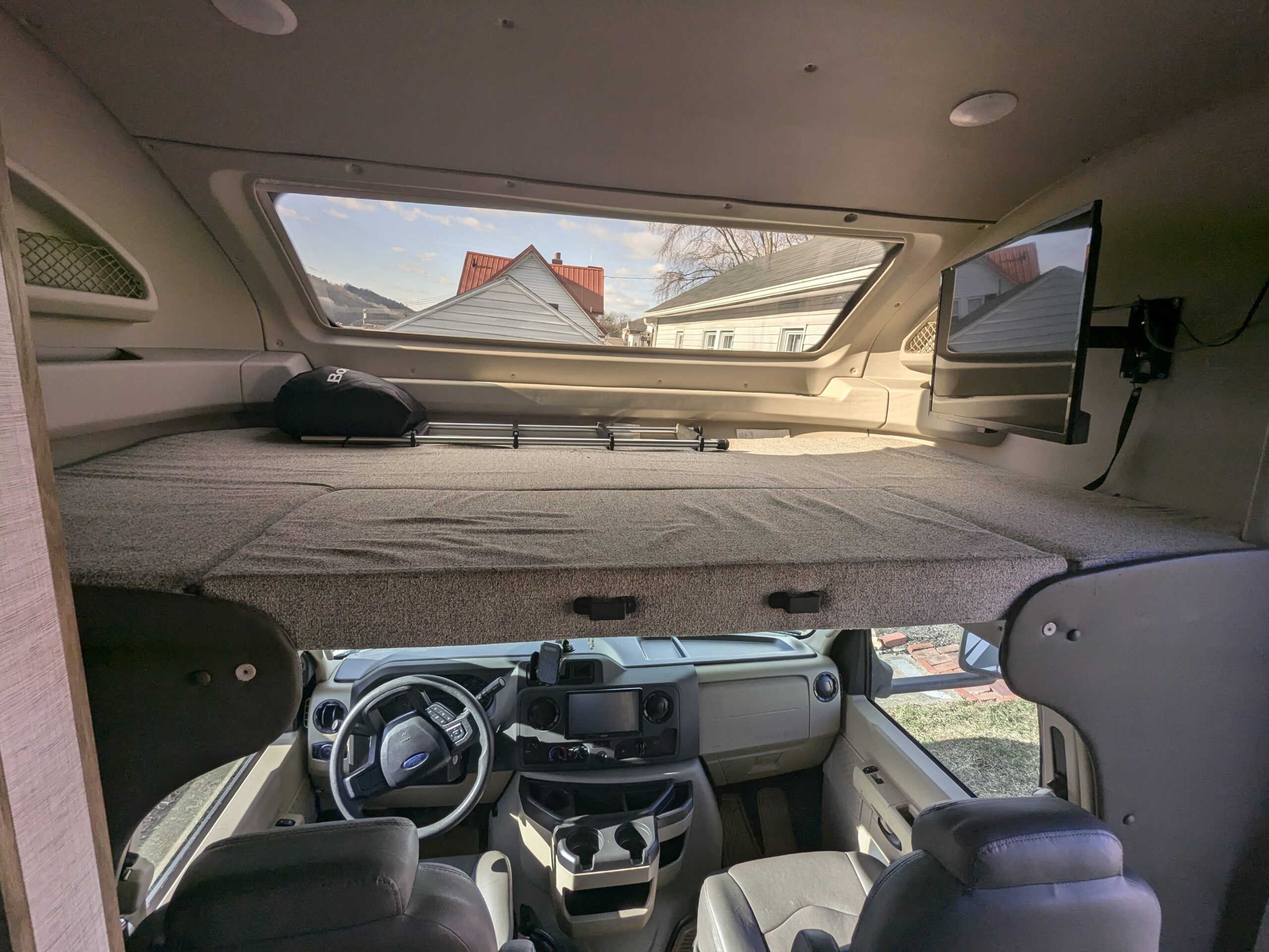 Spacious rv rental interior showing the overhead bunk bed, driver's area, and mounted TV. Houses visible through the large front window.. Entegra Odyssey 2021