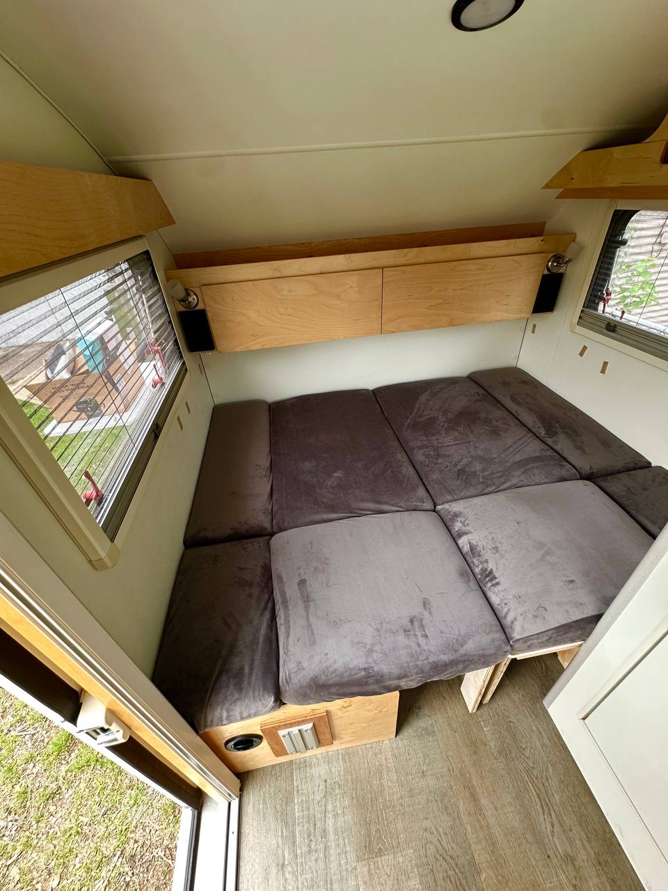 Inside a comfortable RV rental: A plush grey cushioned bed/seating area, overhead wood storage, and bright windows.. nuCamp T@B 2022