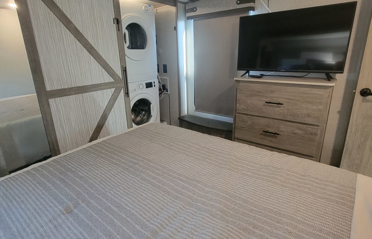 RV rental bedroom featuring a bed, stacked washer/dryer, TV, and dresser, offering convenient compact living.. Forest River Wildwood Heritage Glen 2021
