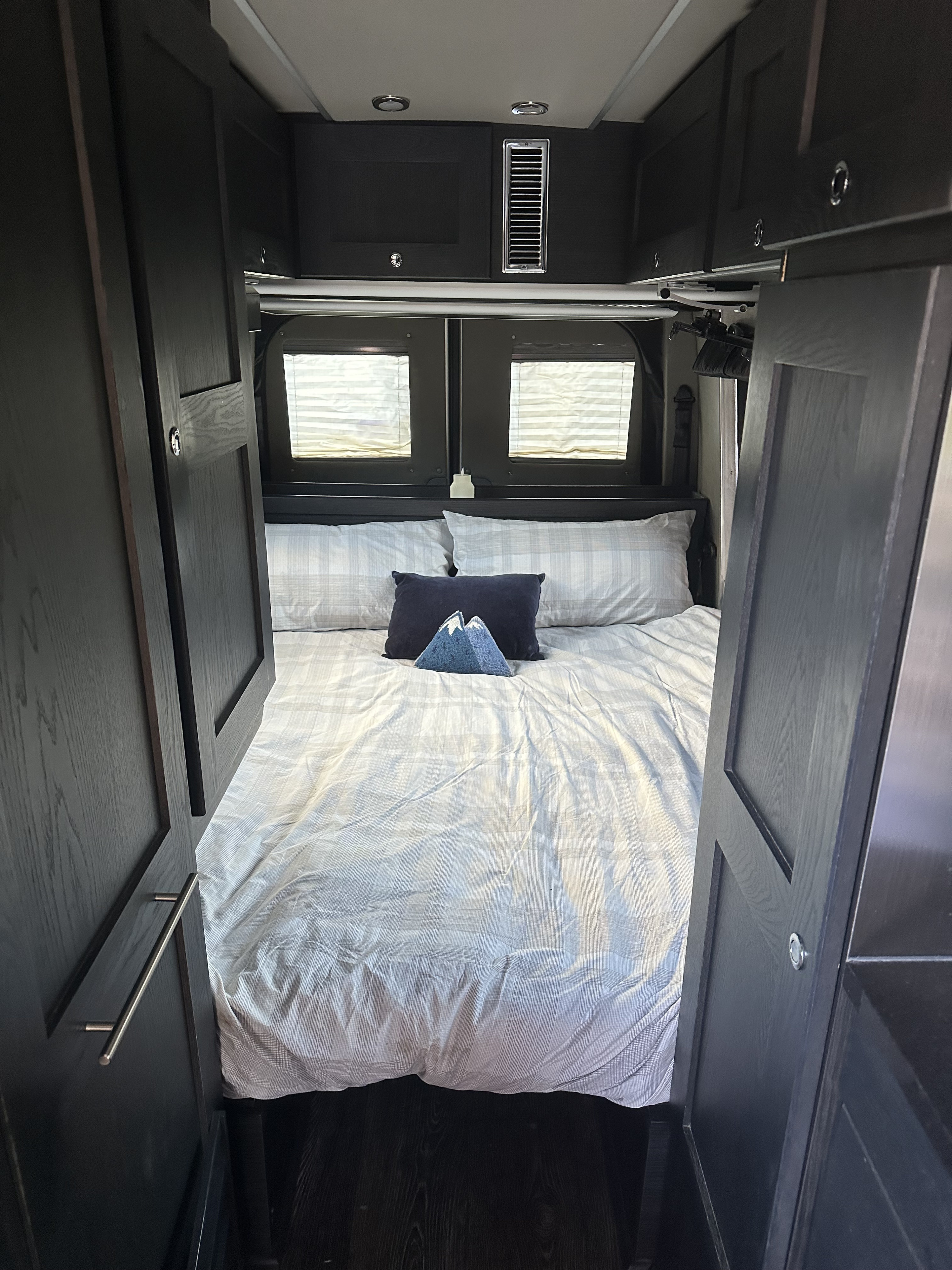 Step into this comfortable RV rental bedroom. Features a large bed with striped bedding, dark cabinets, and rear windows.. Roadtrek Roadtrek Class B 2019