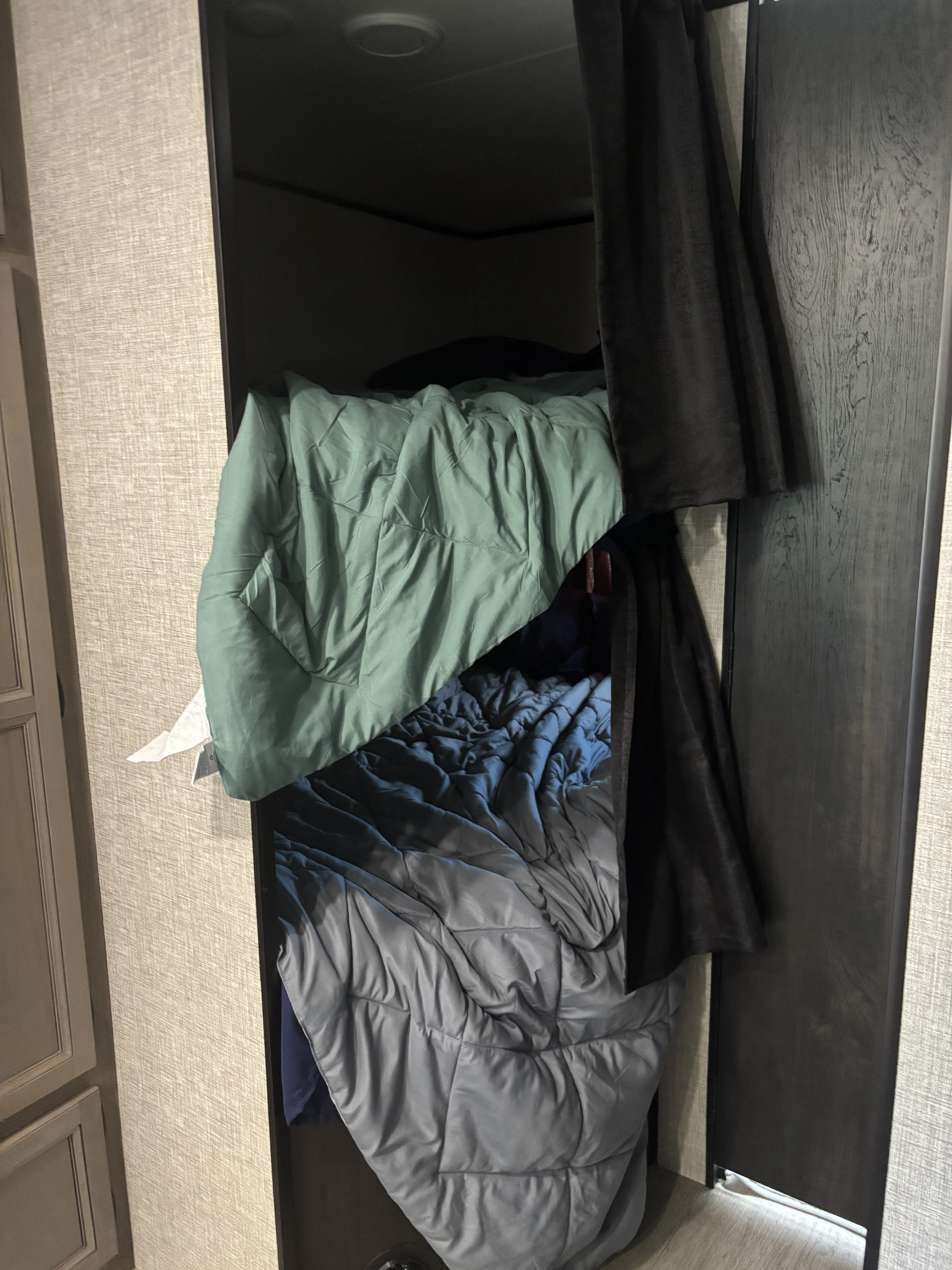 An RV rental's cozy bunk bed, layered with green, blue, and gray comforters. Dark curtains add privacy to the sleeping space.. Jayco Jay Flight SLX 2022