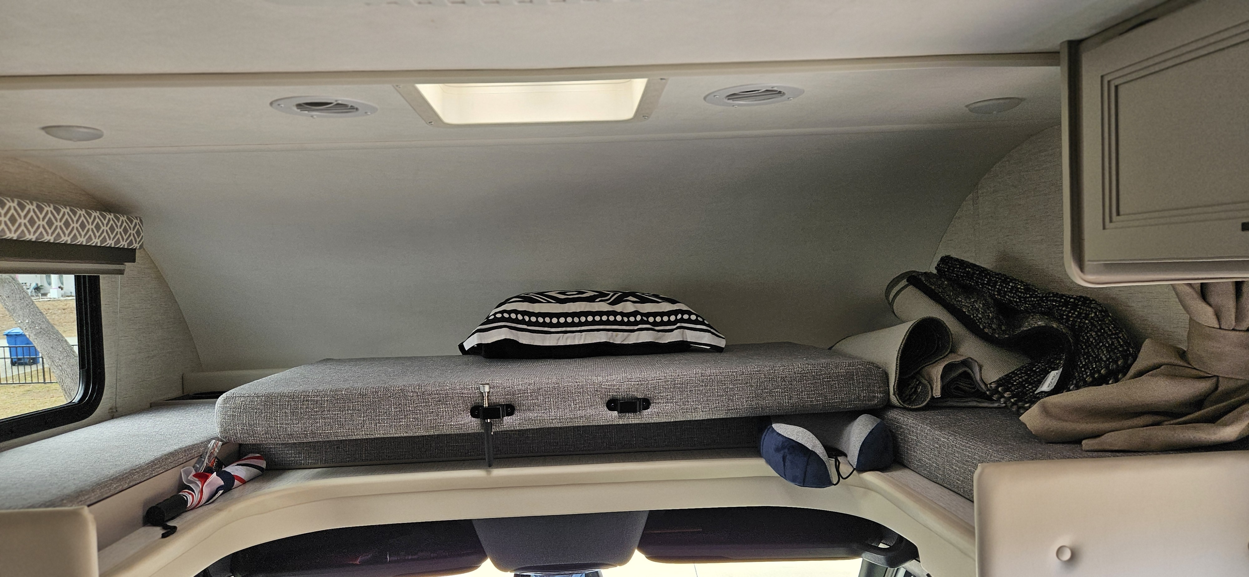Inside an RV rental: a neat overhead bunk with gray cushions, a black/white pillow, and side window. Ready for travel!. Mercedes Coleman 24CL 2024