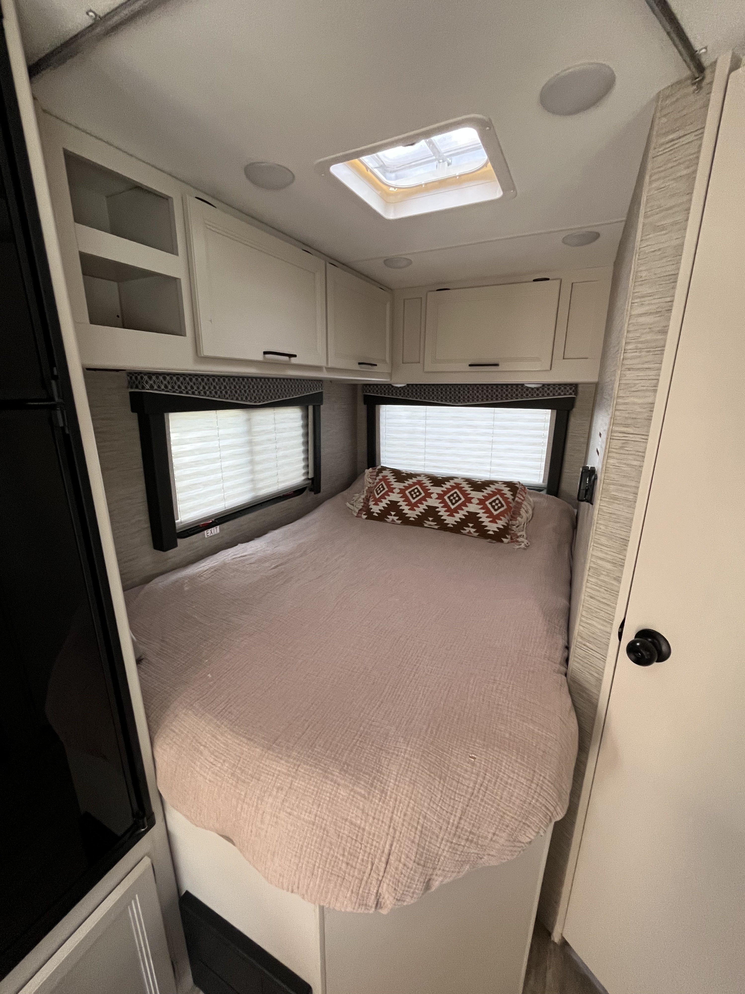 An inviting RV rental bedroom featuring a bed with a patterned pillow, two windows with blinds, and white overhead cabinets.. Thor Chateau 2023