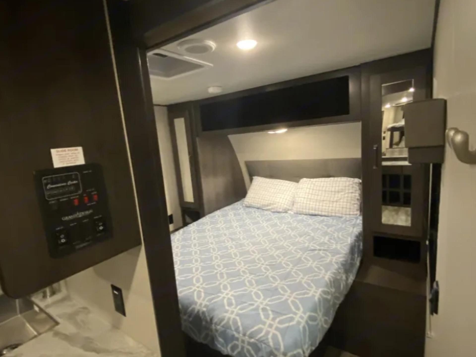 Inside a compact RV rental bedroom, featuring a comfy bed with a blue patterned comforter, overhead storage, and control panel.. Grand Design Transcend Xplor 2021
