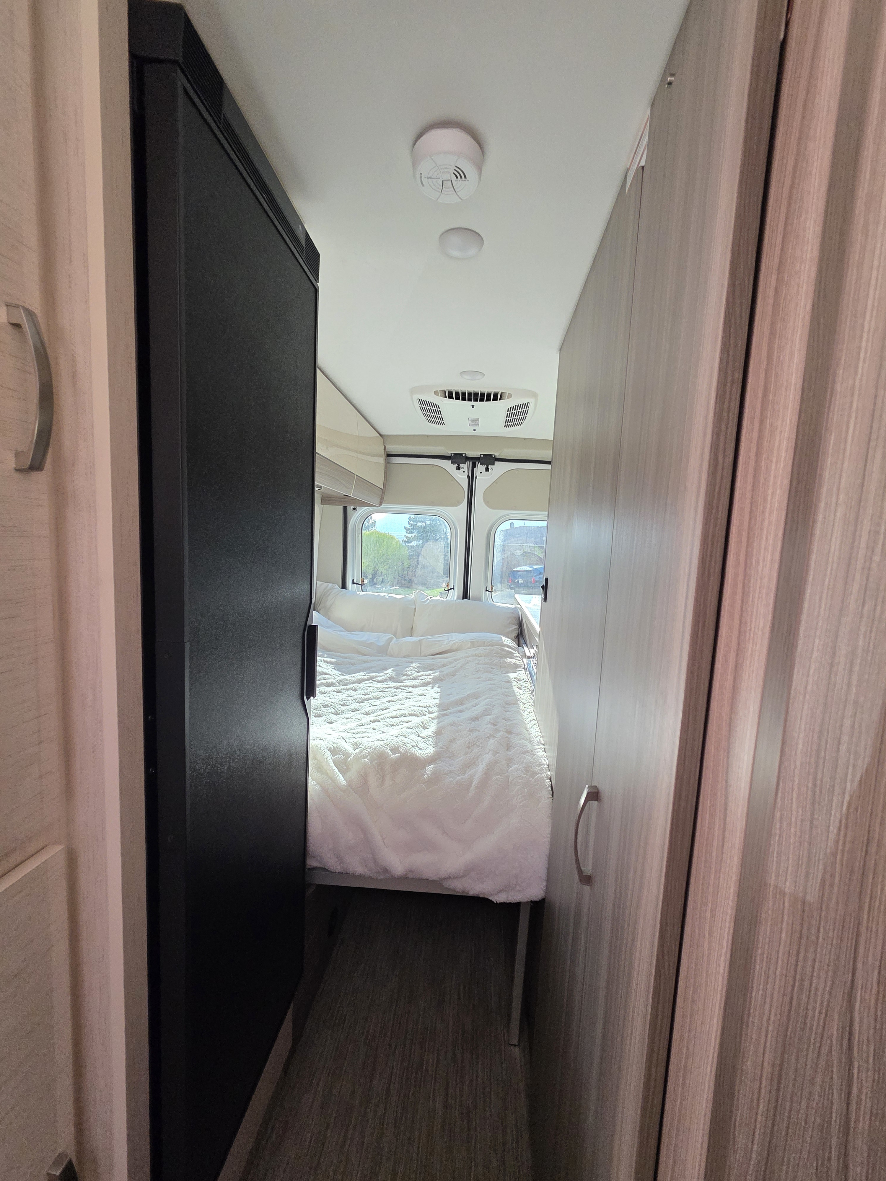 View down a narrow RV rental interior showing a cozy white bed at the end, flanked by a refrigerator and wood-grain cabinets.. Jayco Swift 2024