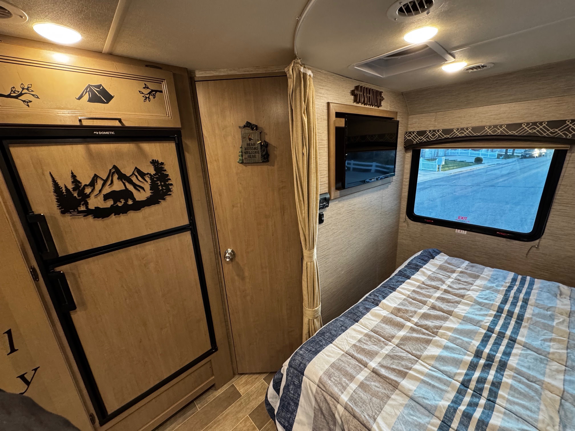 RV rental's cozy interior: striped bed, street view window, Dometic fridge with bear/mountain decor, and wall-mounted TV.. Thor Motor Coach Chateau 2021