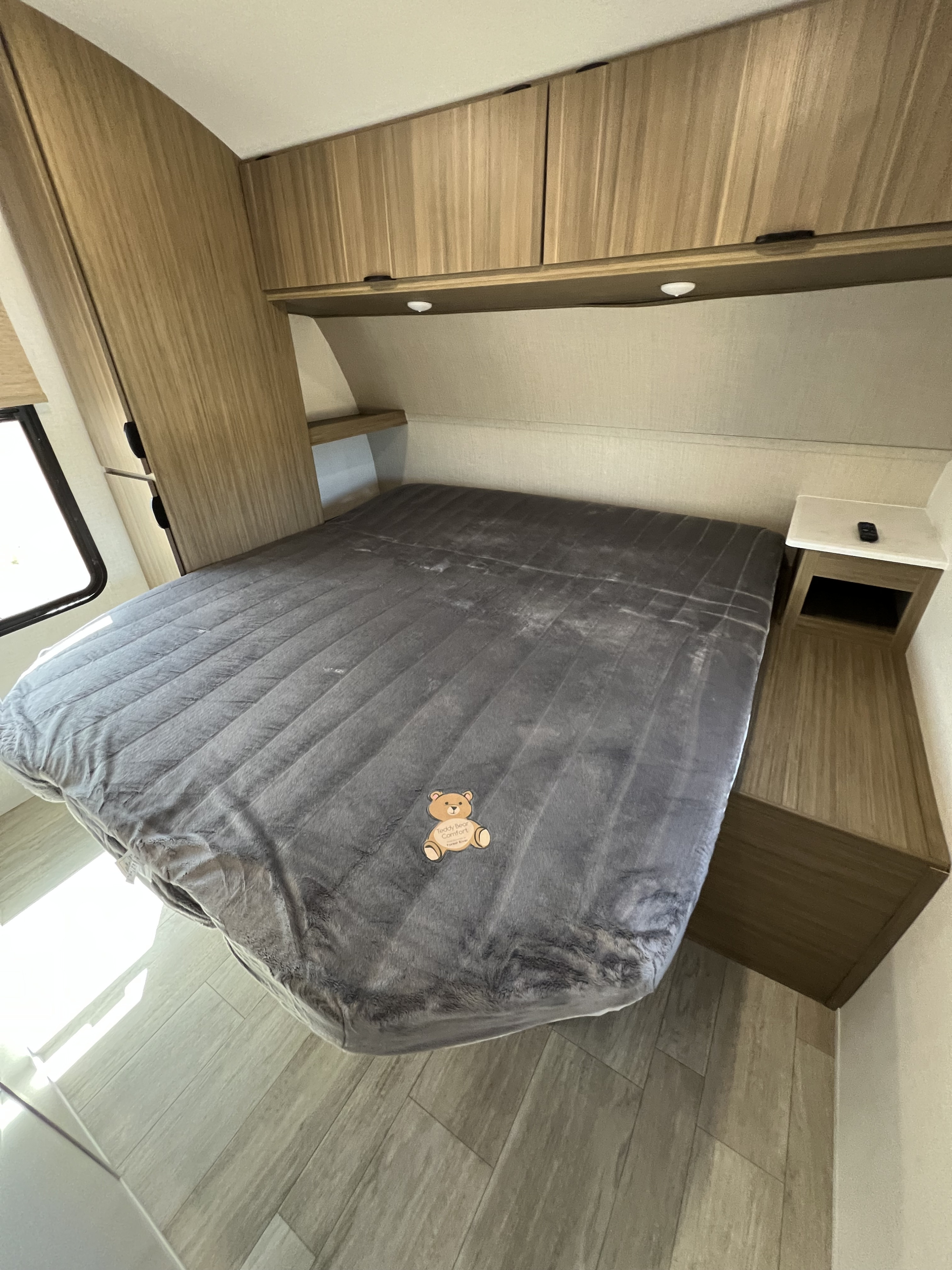 Bright RV rental bedroom with comfy grey bed, wood-look overhead cabinets, and a nightstand. Ideal for travel.. Forest River Salem Cruise Lite 2025