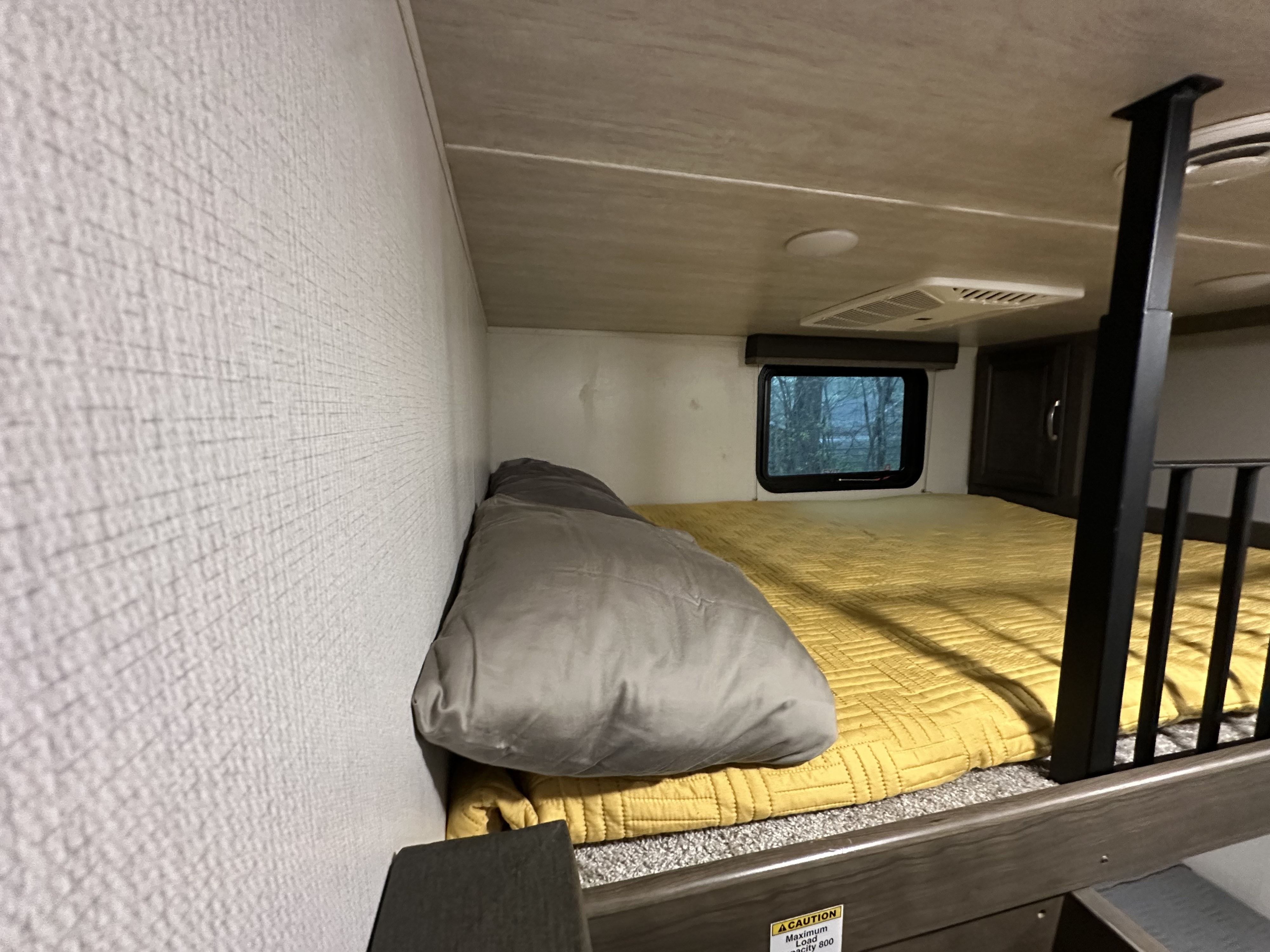 Cozy RV rental bunk bed with a yellow comforter, grey pillow, and window view. Includes AC vent, offering a compact sleeping space.. Keystone RV Cougar 2021