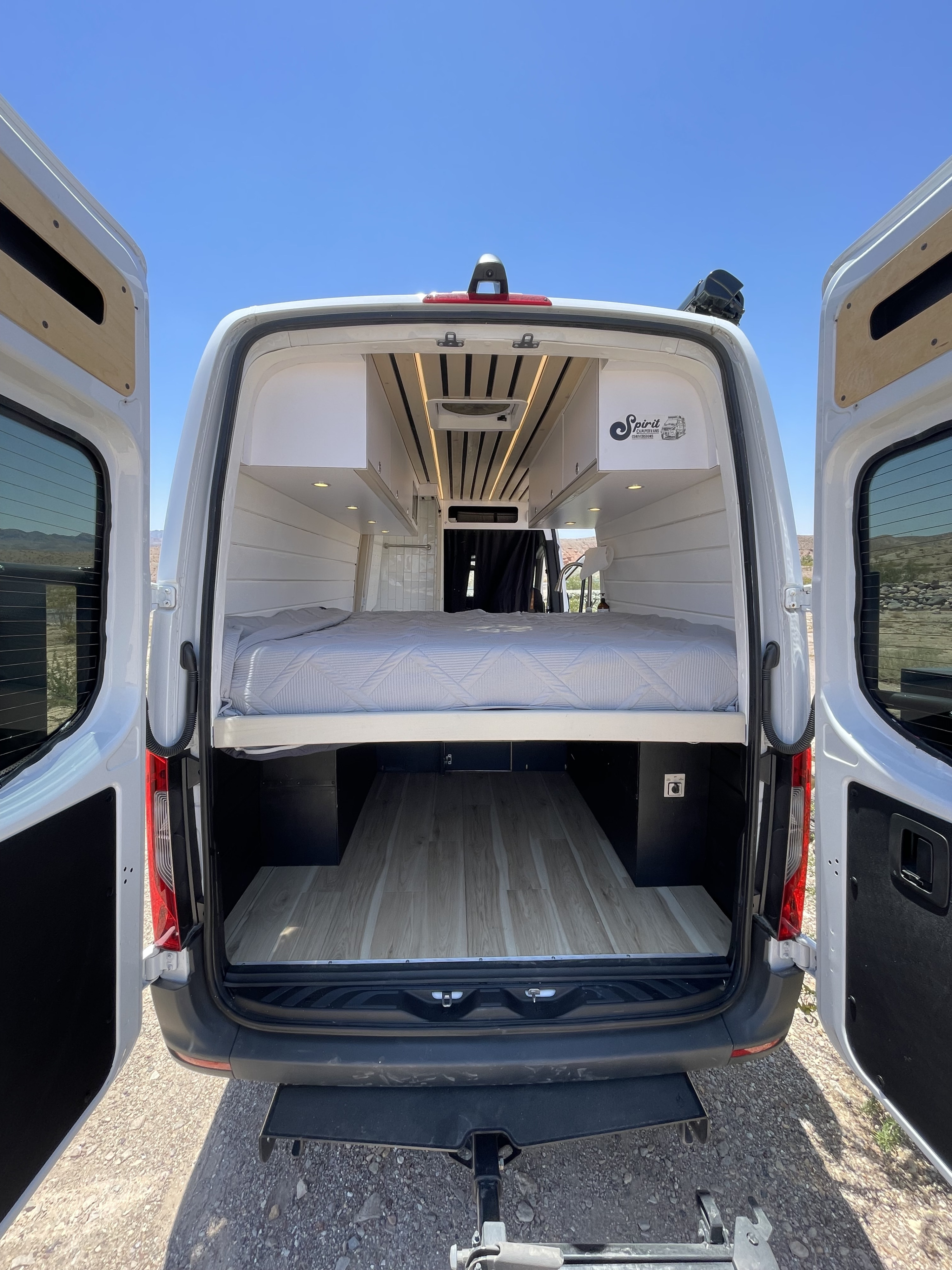 Rear view of a pristine Spirit Campervan Conversions interior with raised bed, large storage, and open doors. Ideal for RV rental.. Mercedes-Benz Sprinter 2021