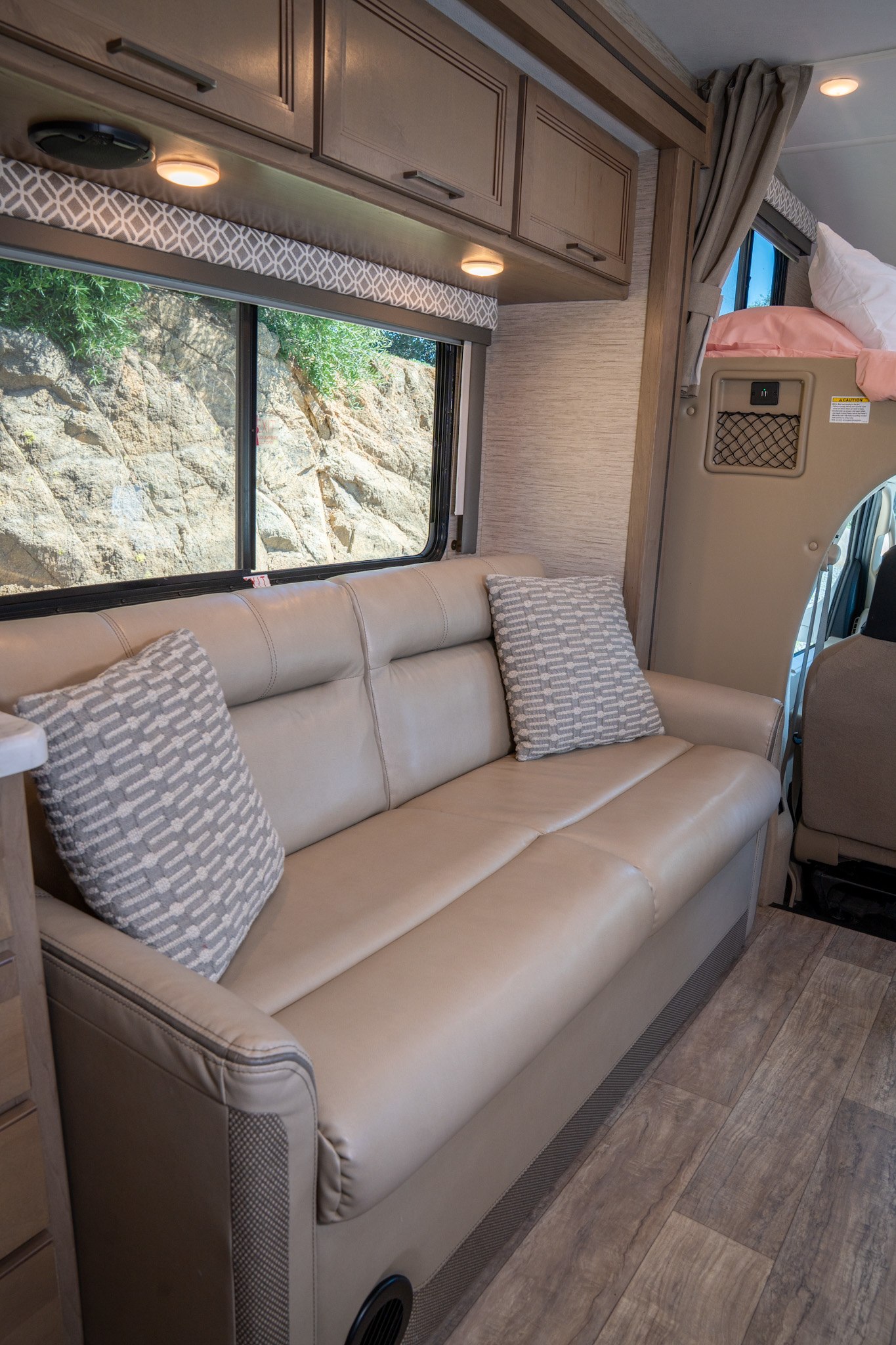RV rental's inviting interior: beige sofa with patterned pillows, wood-look floor, and window revealing a rocky landscape.. Thor Motor Coach Coleman 2023