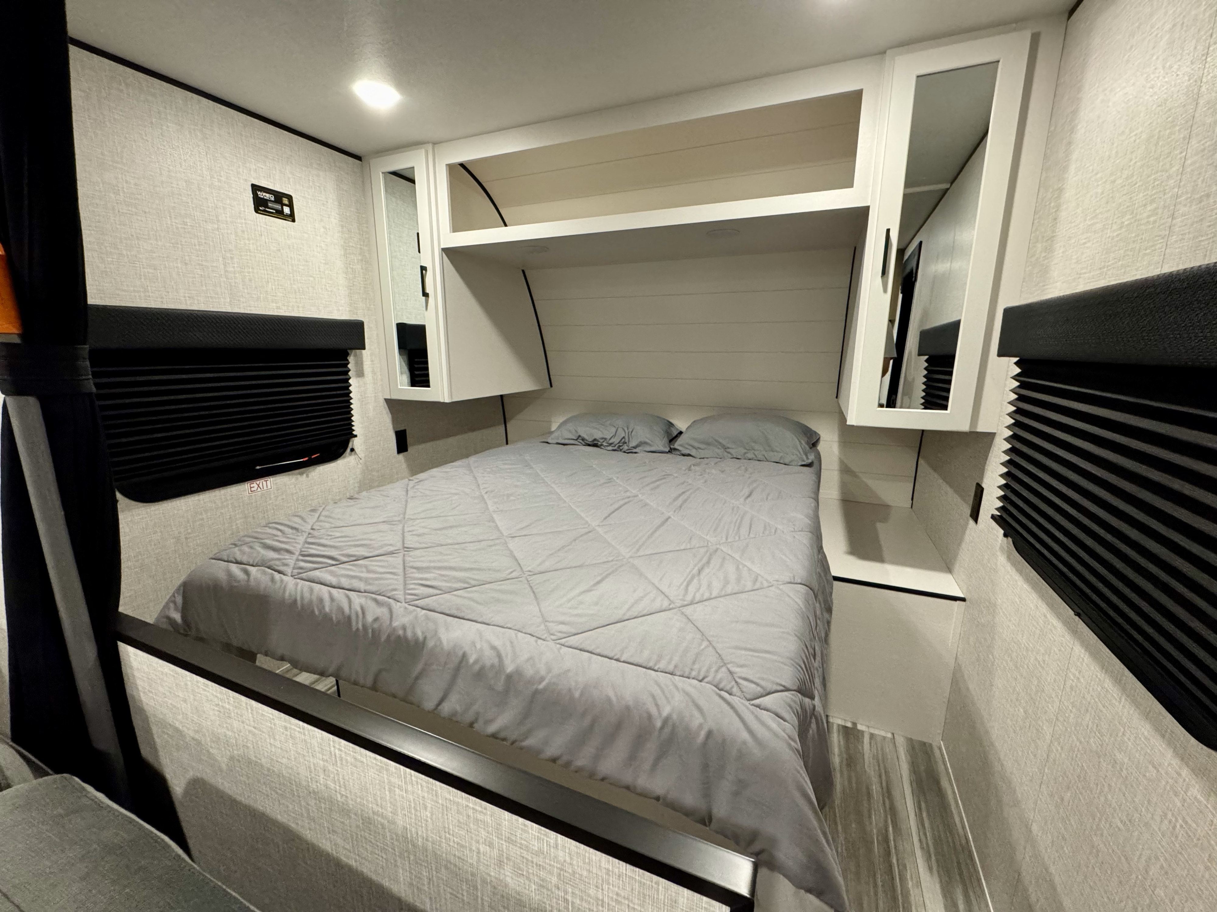 Inside an RV rental: a neat bedroom with a grey bed, overhead storage, and blackout blinds for a good night's sleep on the road.. Jayco 260BH 2025