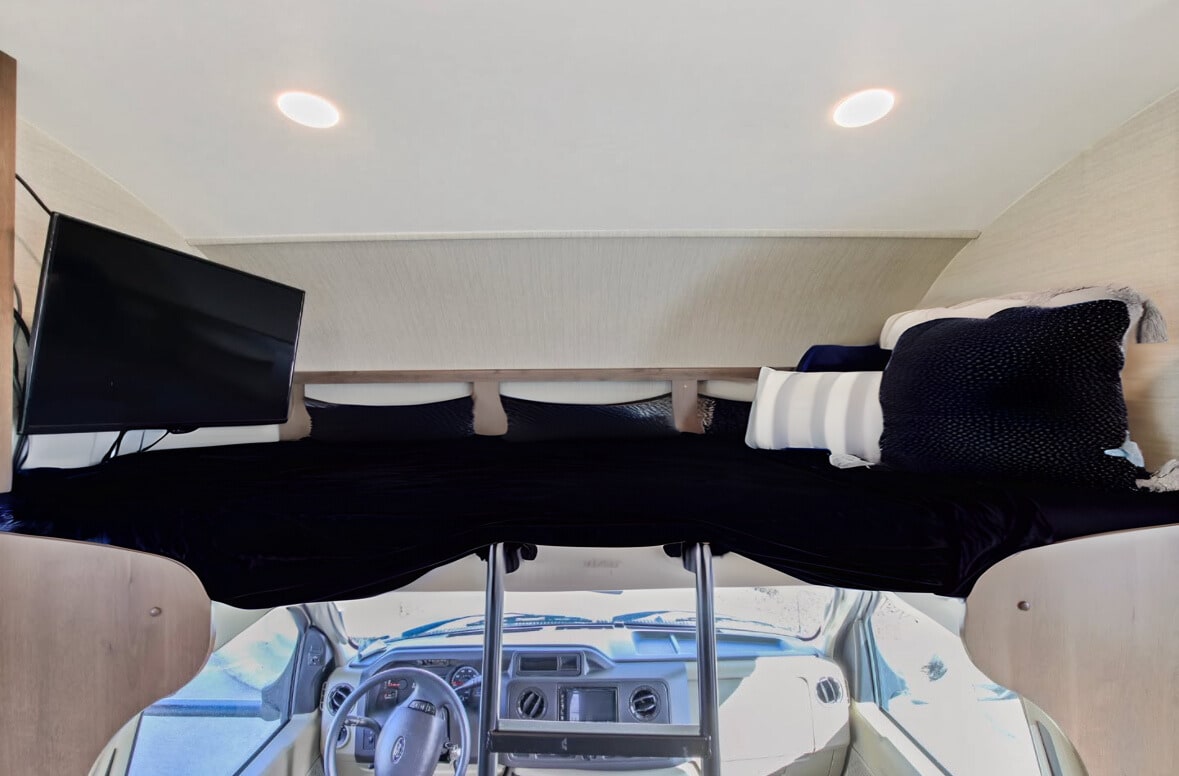 Cozy RV rental cab-over bunk with dark bedding, pillows, TV on the left, and the vehicle's dashboard visible below.. Jayco Redhawk 2019