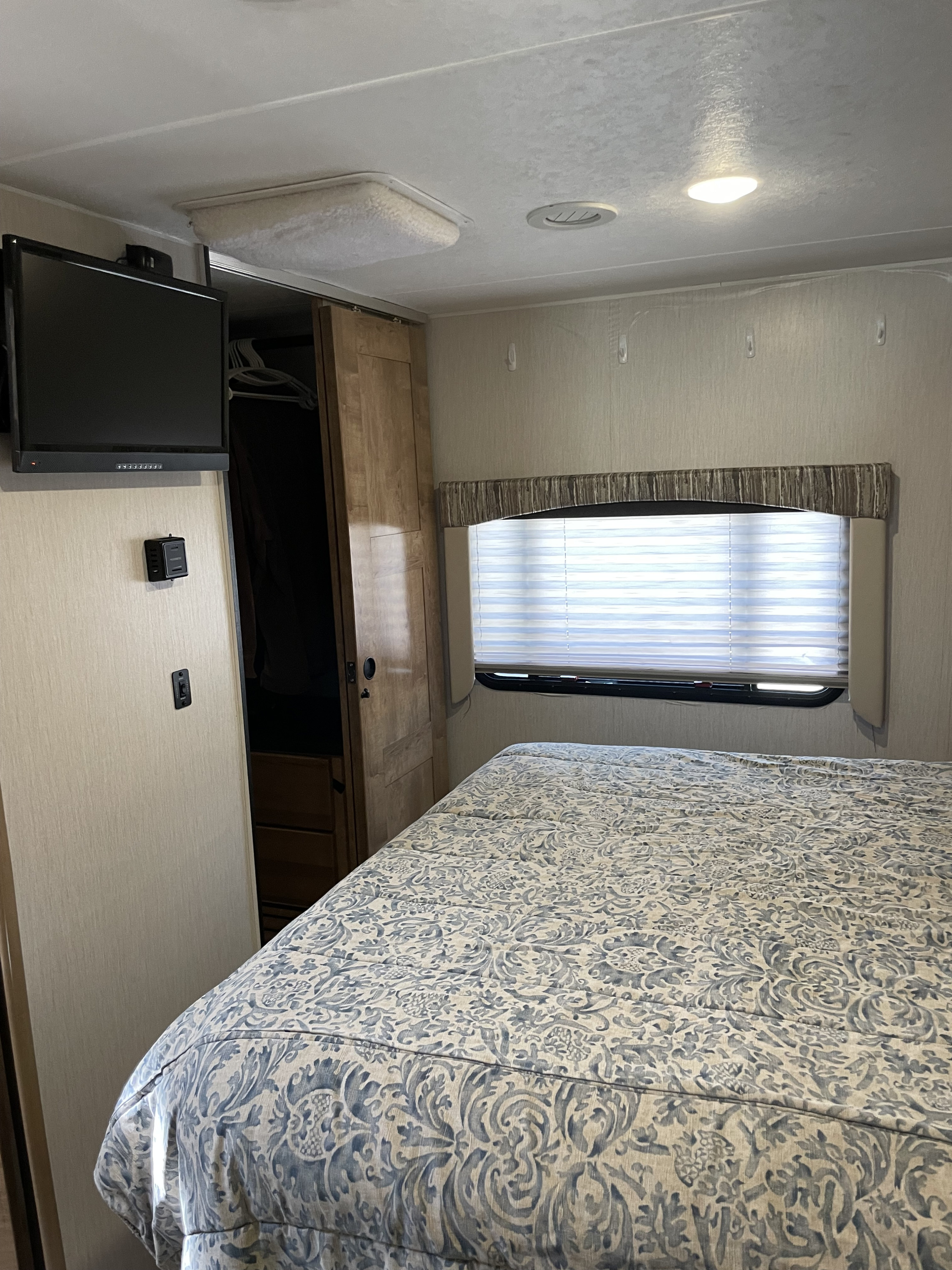 This RV rental bedroom features a patterned bed, wall-mounted TV, and a window with blinds, showing a compact but functional space.. Forest River Coachmen Leprechaun 2020