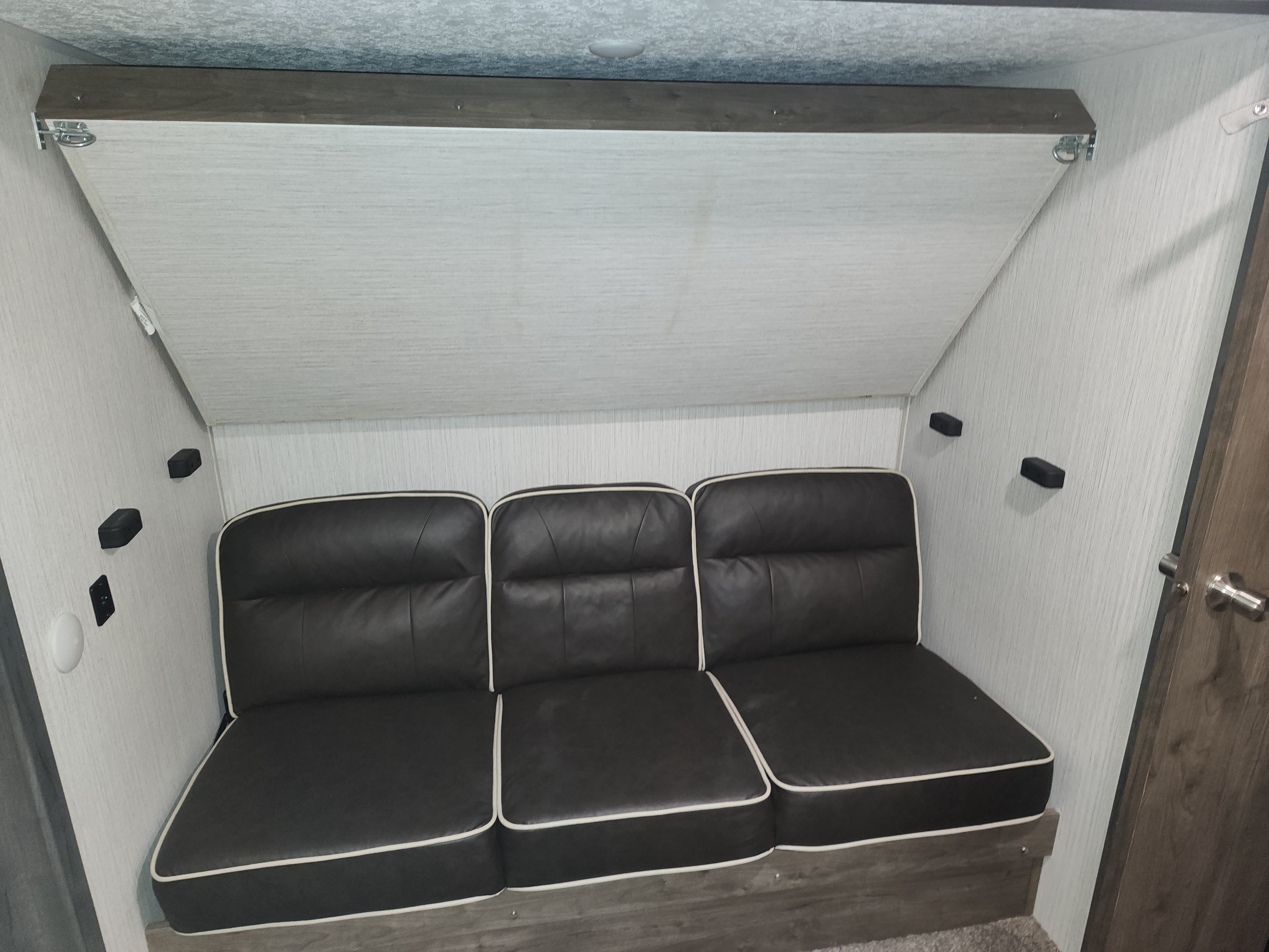 RV rental interior features a dark three-seater sofa and an overhead, secured fold-down bunk bed, optimizing space.. Forest River Puma 2020