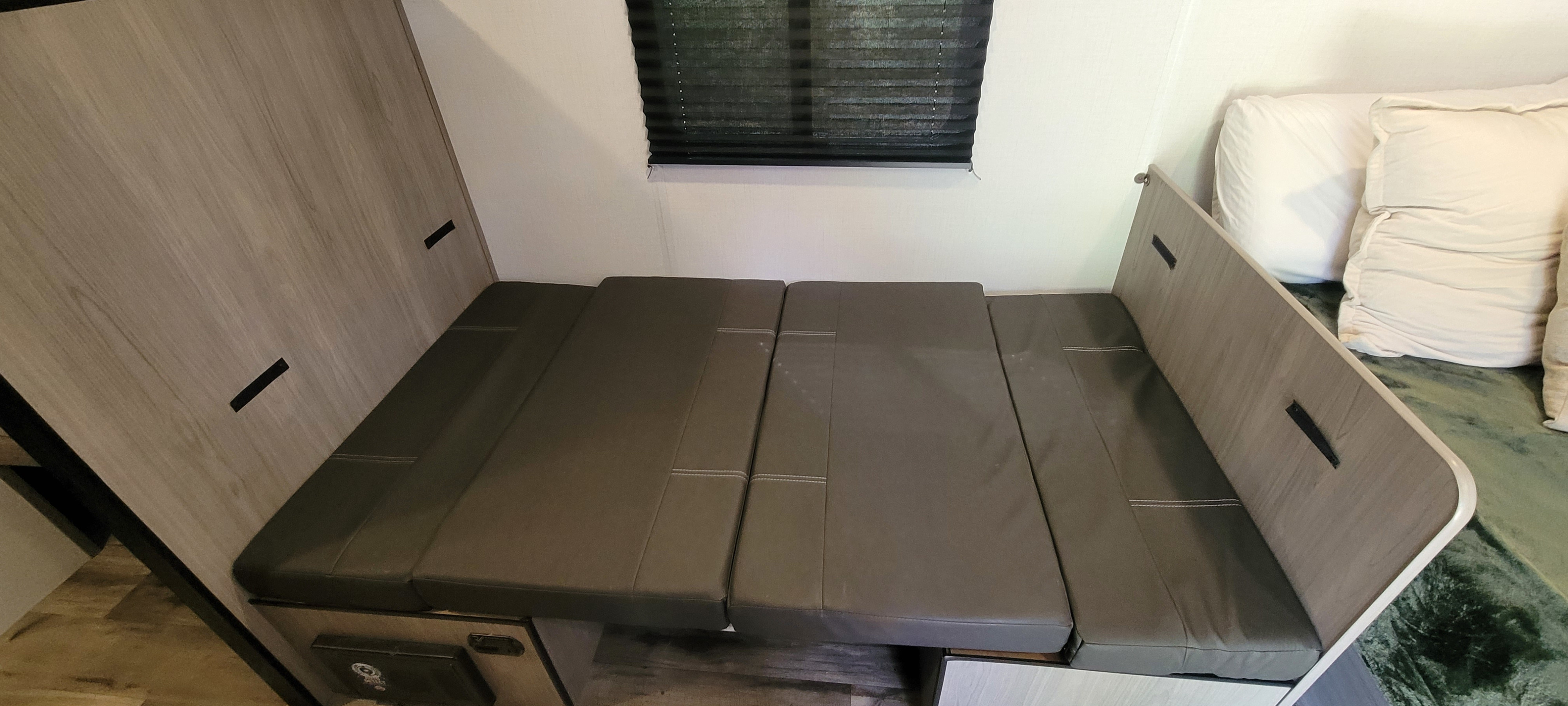 RV rental interior: dinette converted into a bed with gray cushions and a window blind.. Dutchmen 2024 Dutchmen Coleman Lantern 17B 2024