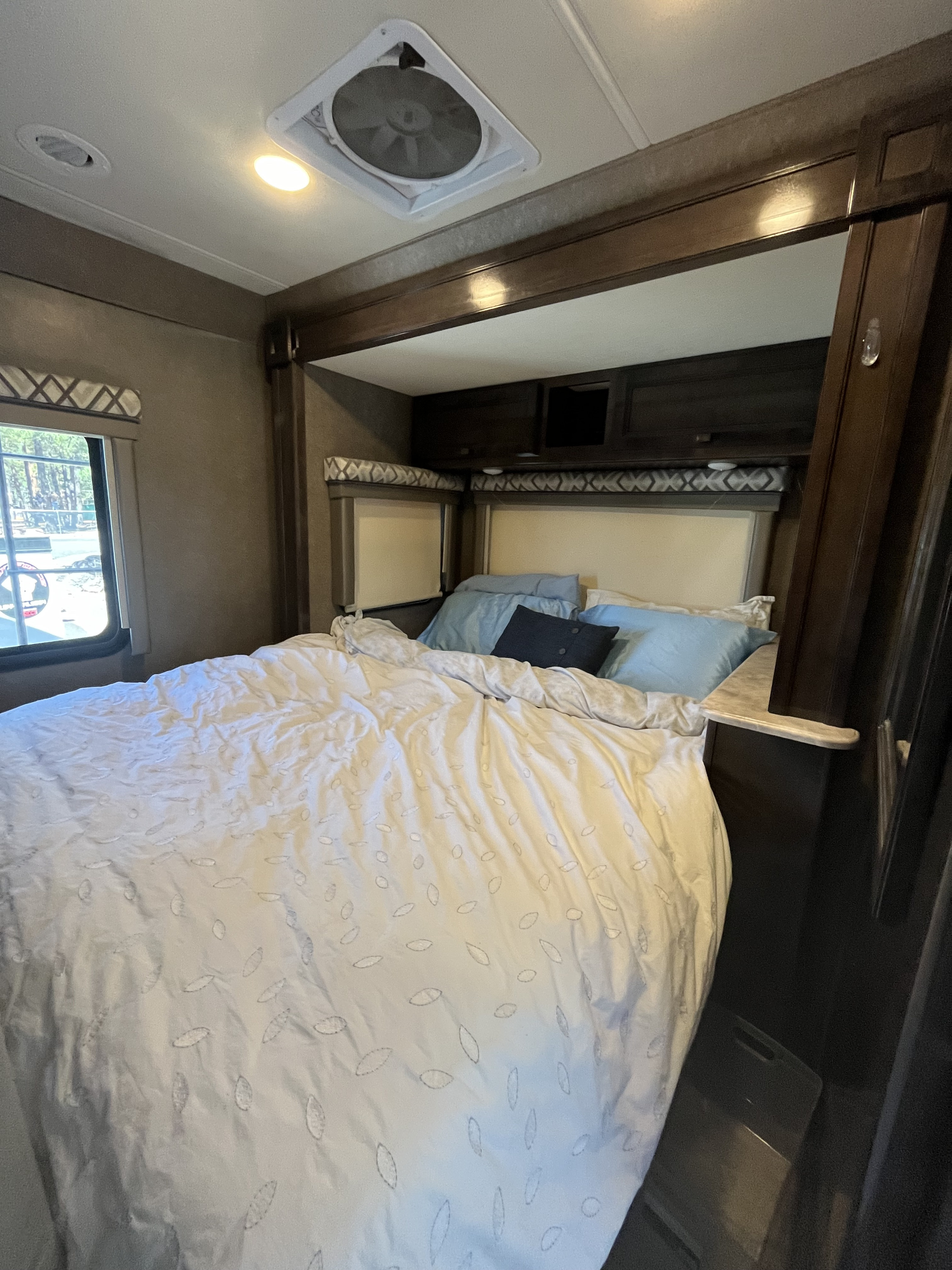 Bright RV rental bedroom featuring a bed with patterned duvet, overhead cabinets, windows, and ceiling vent.. Thor Siesta 2020