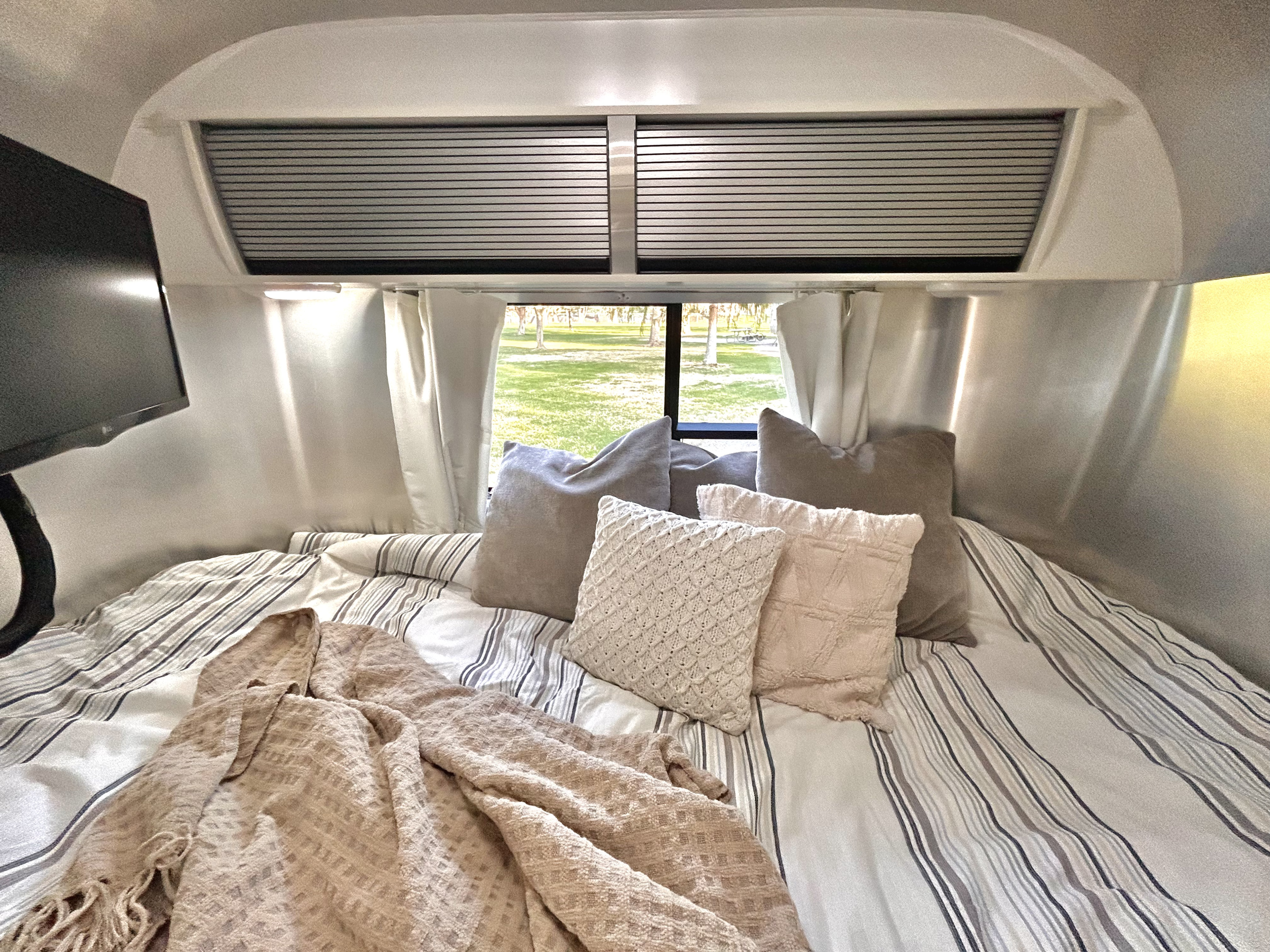 Cozy RV rental bed with striped bedding, various pillows, and a blanket, offering a relaxing view of nature.. Airstream Sport 2018