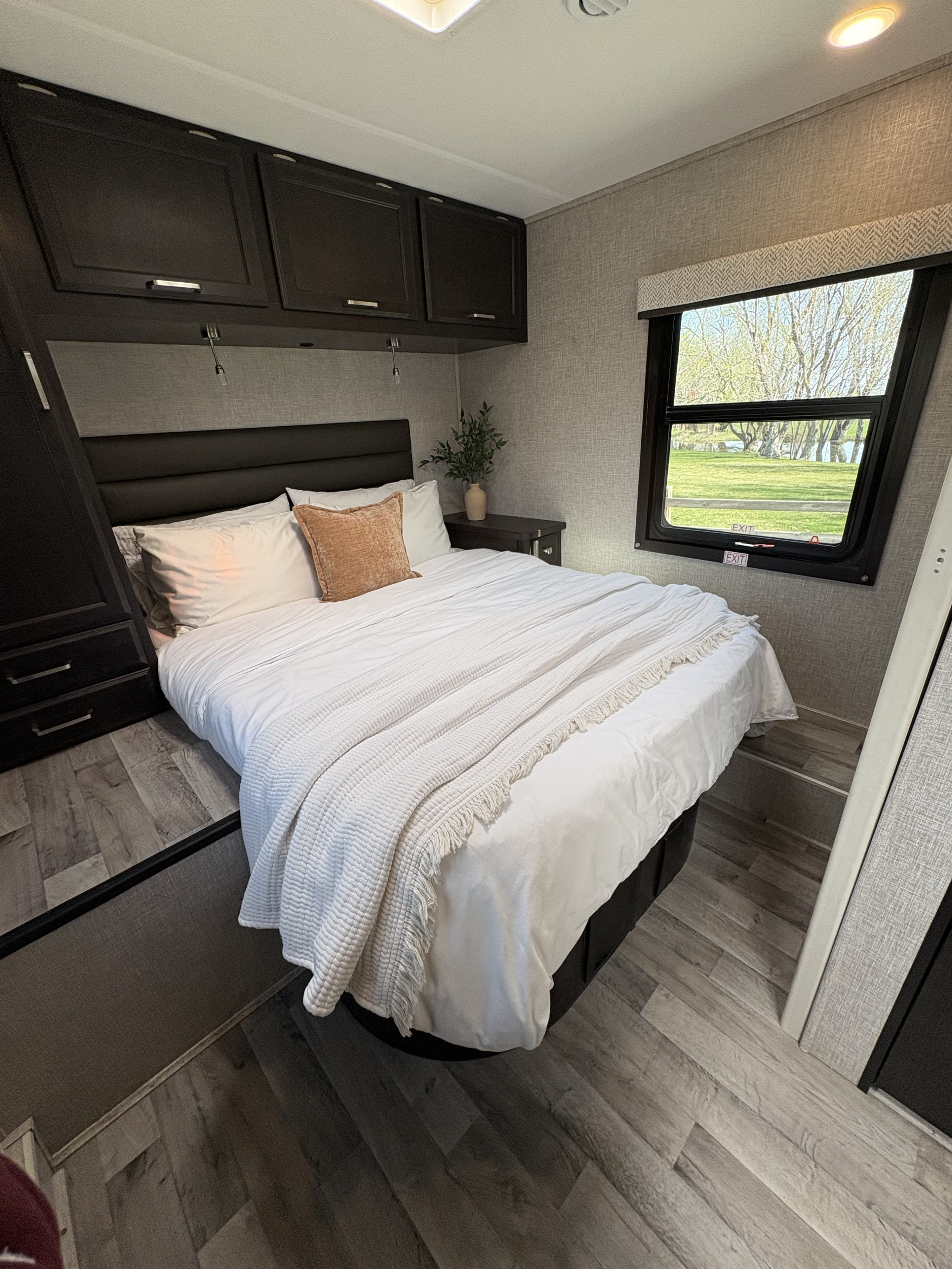 Serene RV rental bedroom: cozy bed with white linens, dark storage, and a scenic window overlooking nature.. Jayco Redhawk 2024