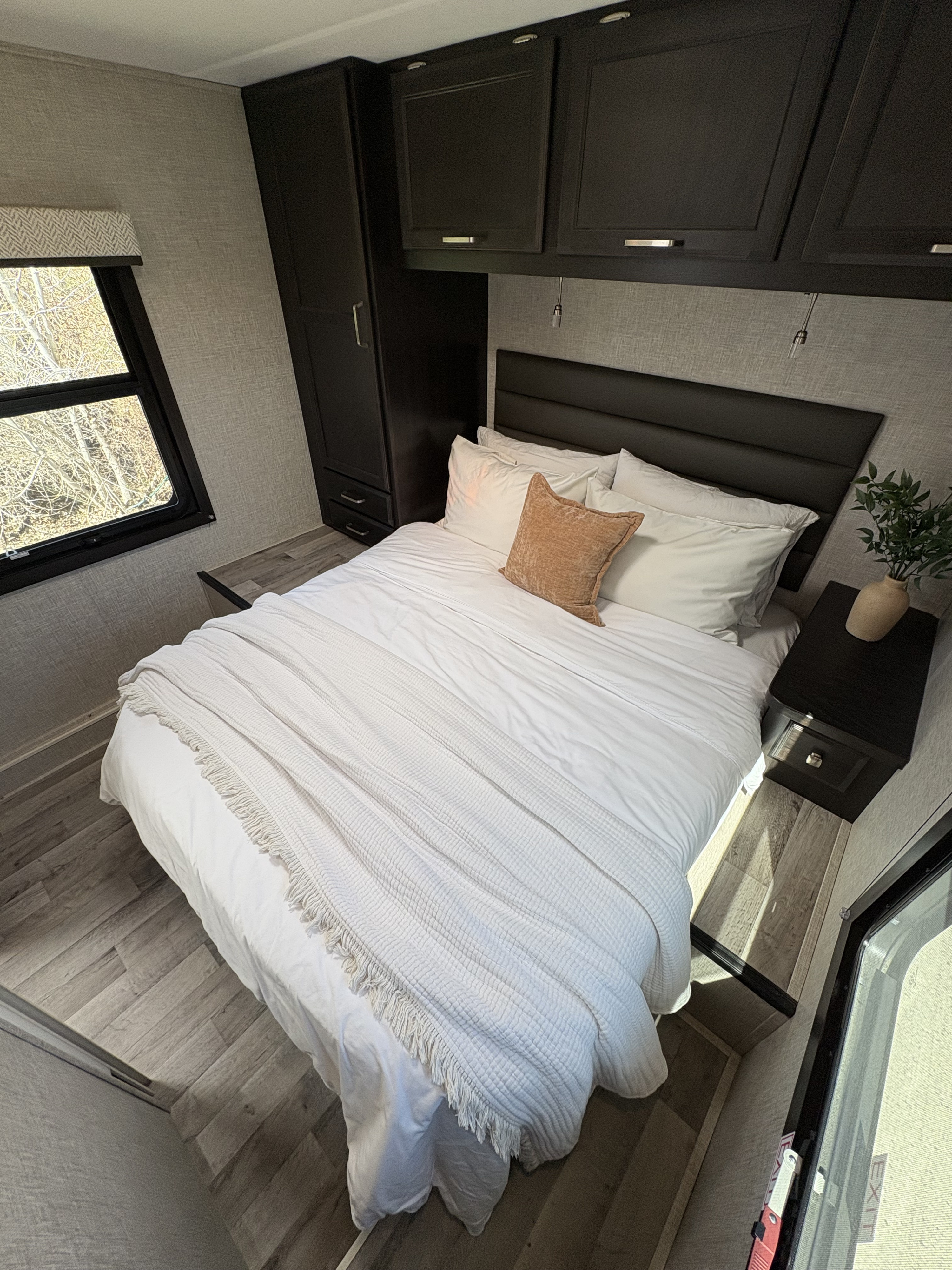 This clean RV rental bedroom offers a comfortable bed with white linens, dark cabinets, and a window view.. Jayco Redhawk 2024