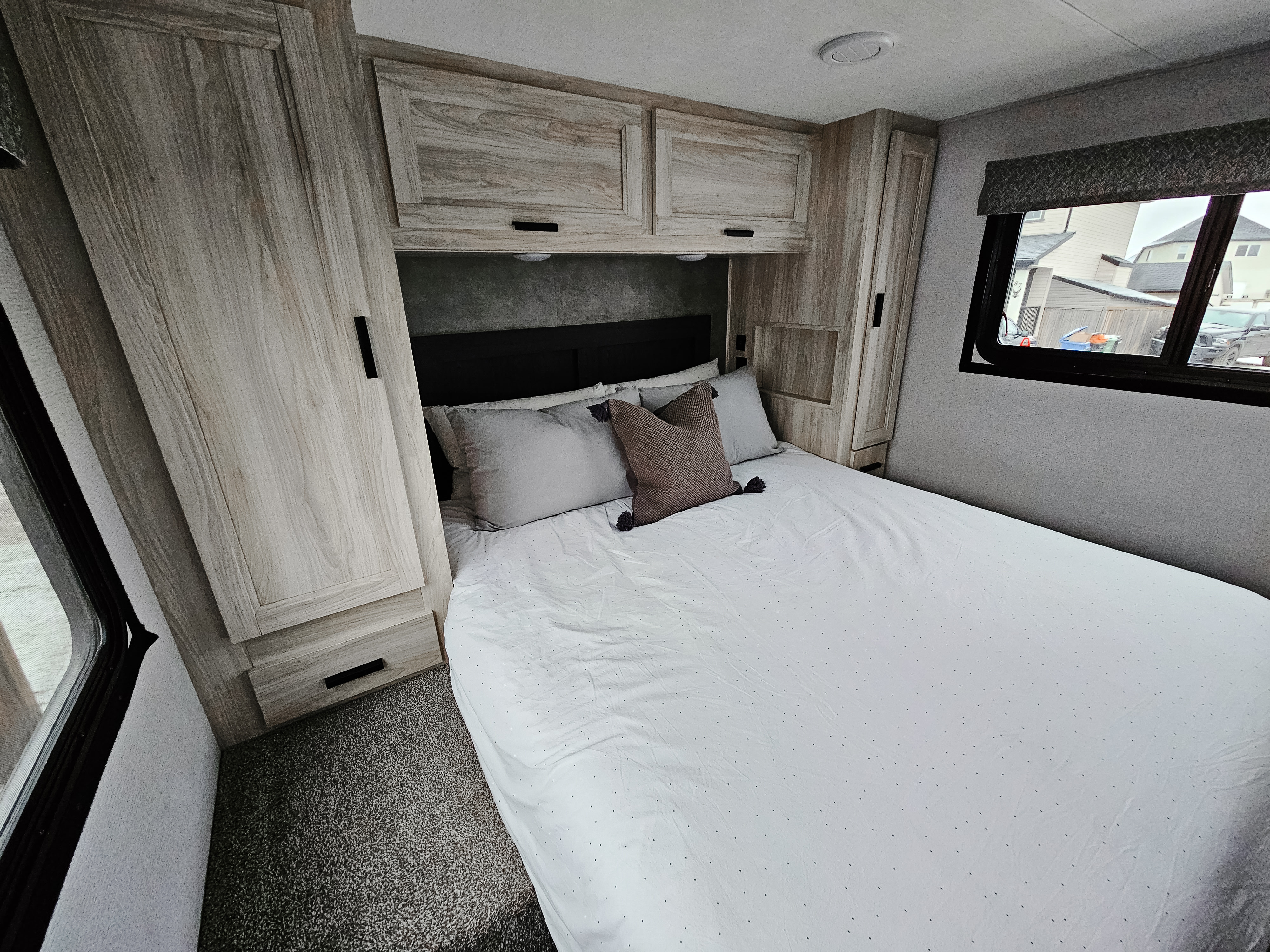 Modern RV rental bedroom with a large bed, light wood cabinets overhead and flanking it, and a window to the right. Grey carpet.. Forester 2651 CDW 2022