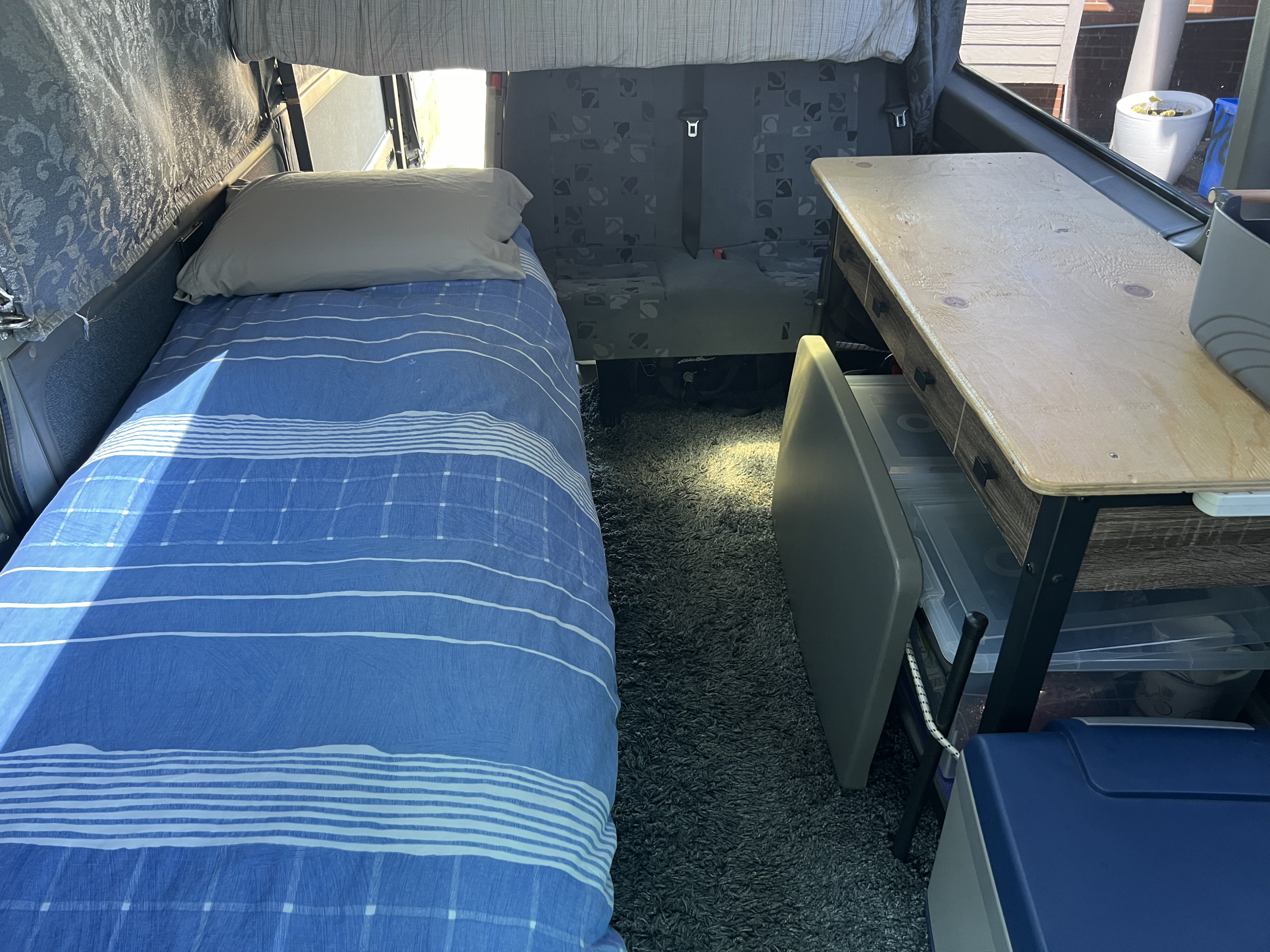 Inside an RV rental: a blue-striped bed on plush carpet, a wooden table with storage bins for comfortable mobile living.. Dodge Sprinter Van 2006