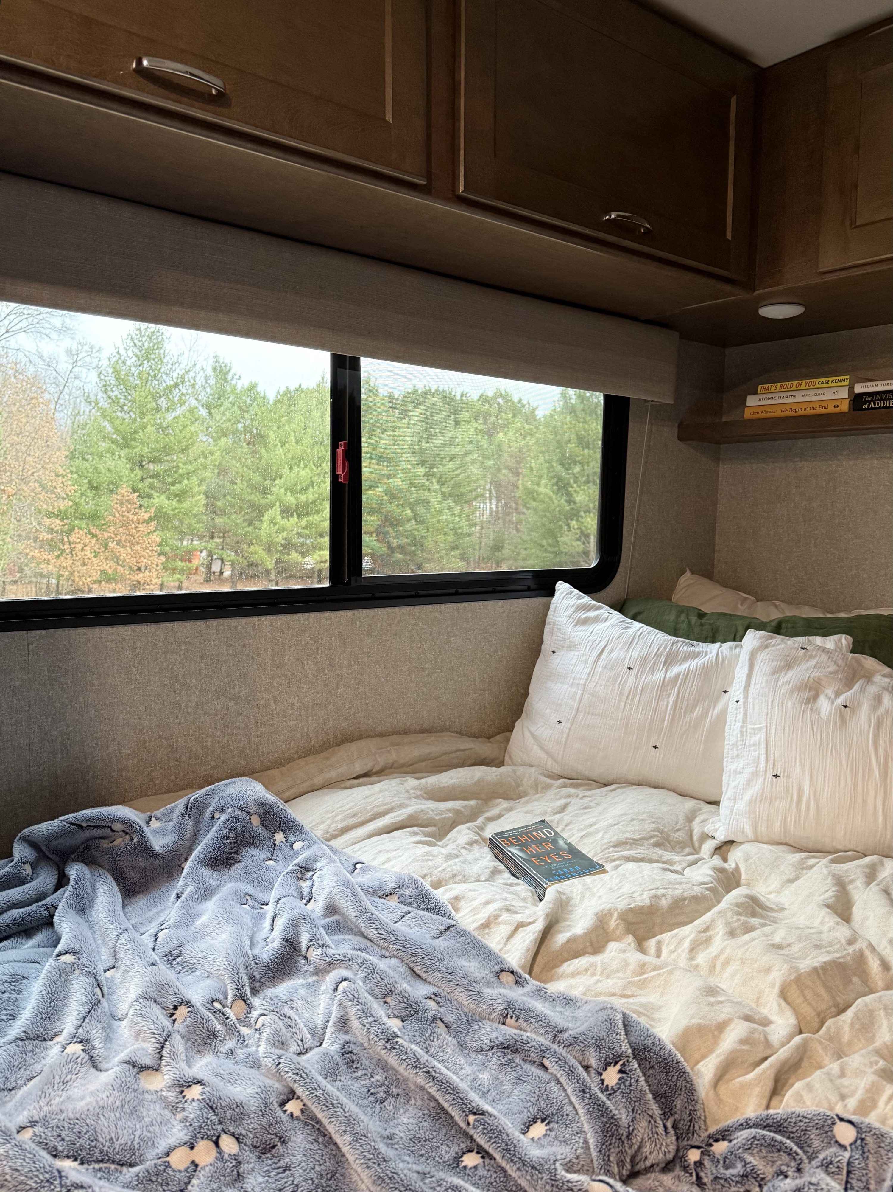 A serene rv rental interior featuring a cozy bed with a book, fluffy blanket, and large windows offering a lush forest view.. Winnebago Vita 2020