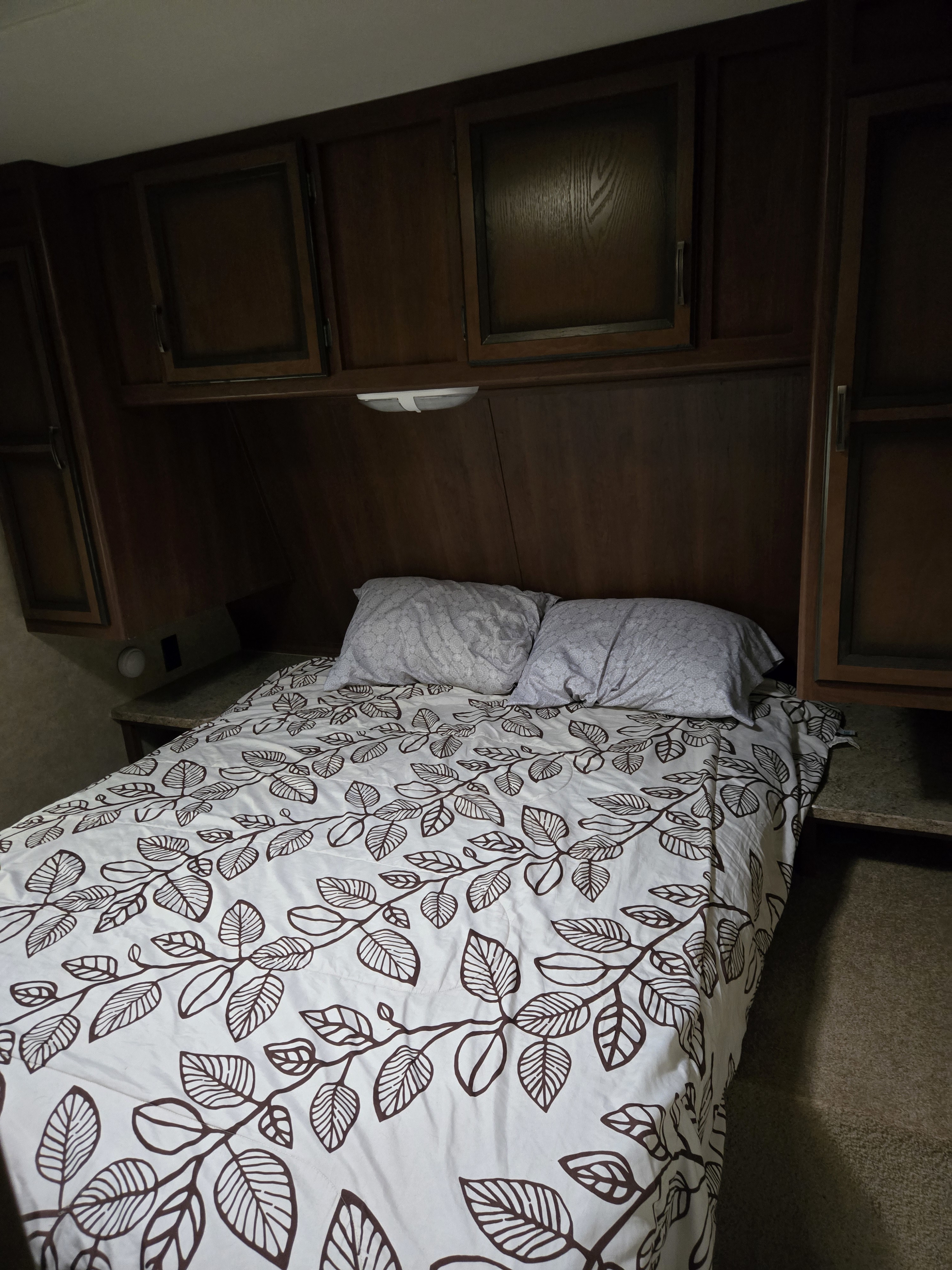 Inside an rv rental: a cozy bedroom with leaf-patterned bedding, two pillows, and dark wood cabinetry overhead.. Crossroads RV Zinger 2014