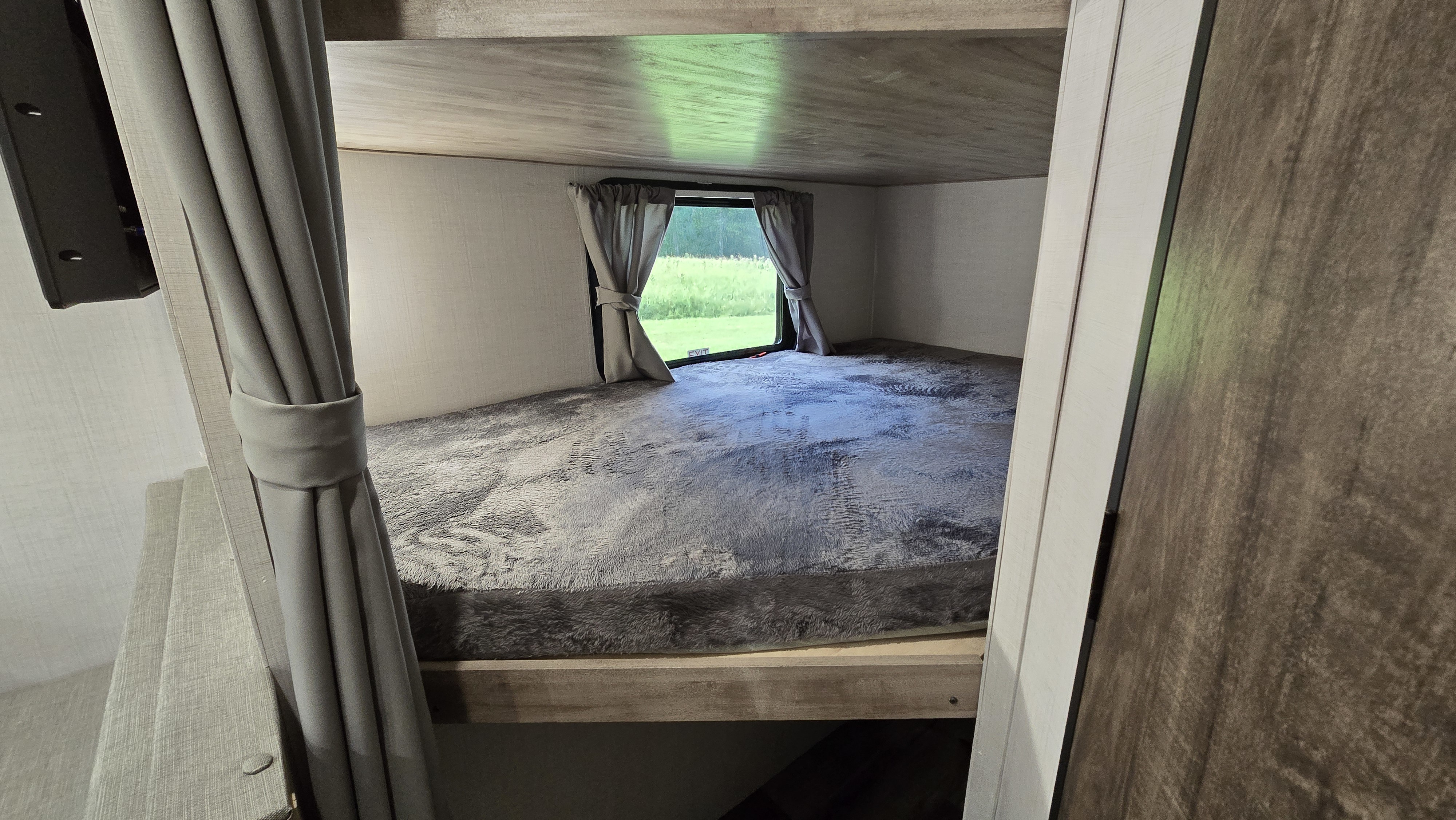 Inside an RV rental, a comfy bunk bed with grey blanket and window overlooking green nature.. Gulf Stream Trailmaster 2024