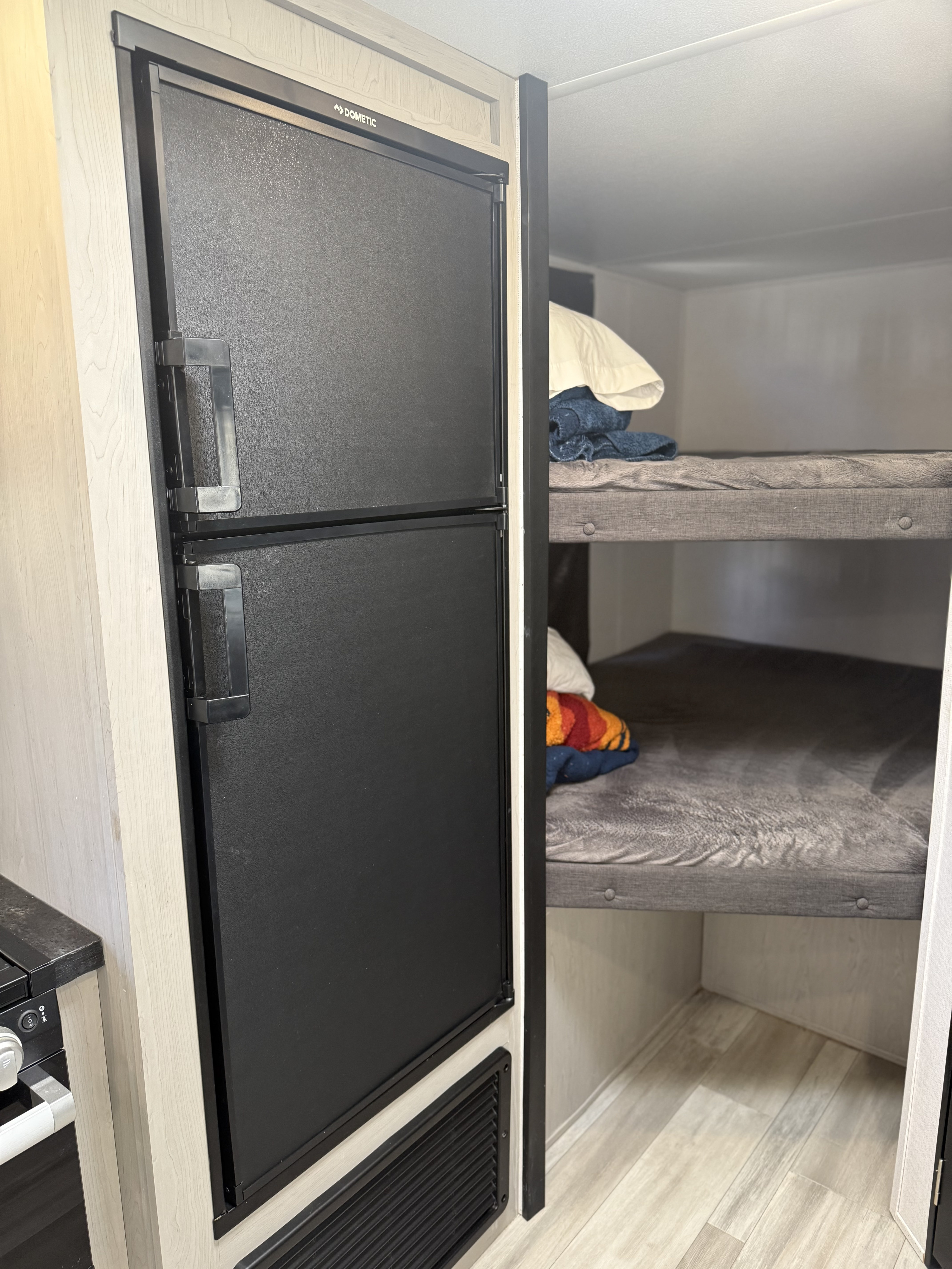 Inside an RV rental: a black Dometic refrigerator and two gray bunk beds with bedding, set against light wood cabinetry.. Primetime Tracer 2022
