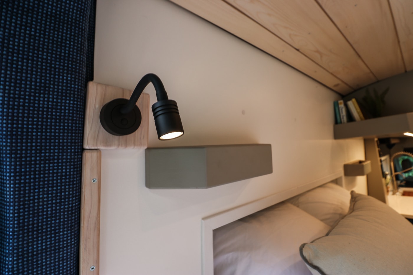 Close-up of an RV rental bedroom showing a black reading lamp, light wood accents, bed, and gray floating shelf.. Ford Transit 2016
