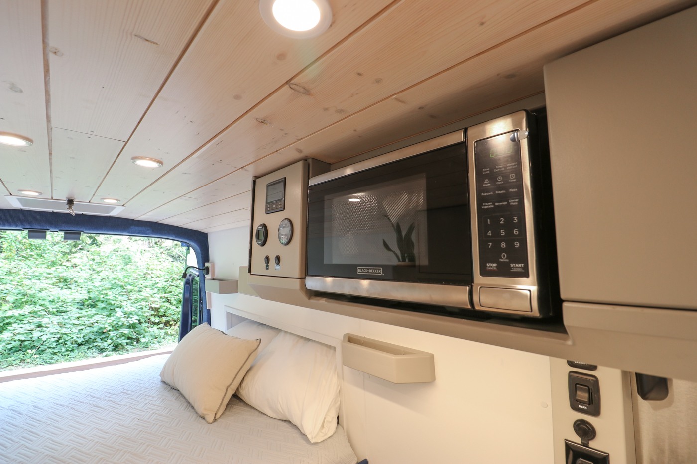 This RV rental interior features a Black+Decker microwave and control panel above a cozy bed, with a view of lush greenery.. Ford Transit 2016