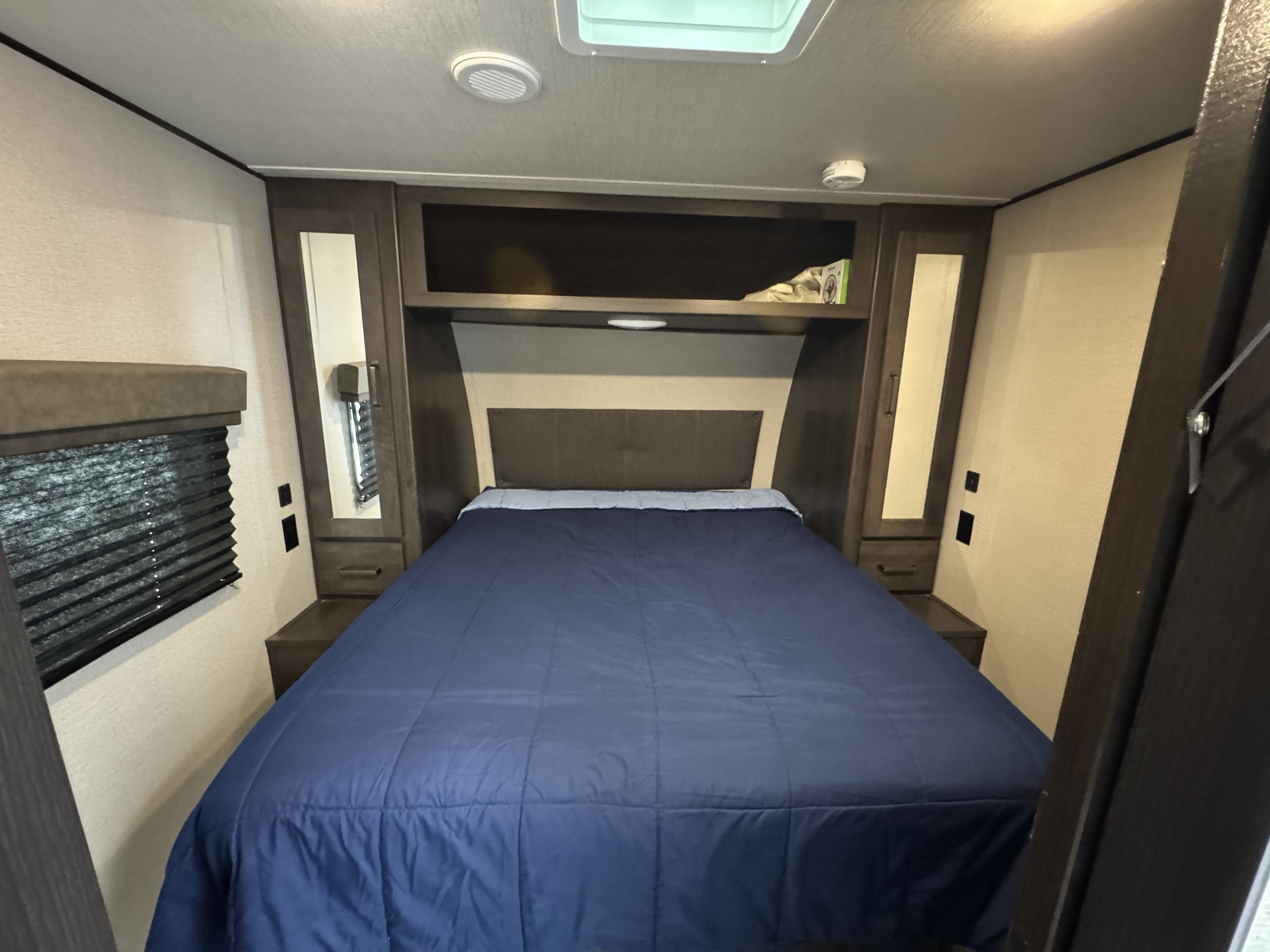 Inside an RV rental, a neat bedroom features a large bed with a blue comforter, built-in storage, and window, ready for your trip.. Grand Design Transcend Xplor 2022