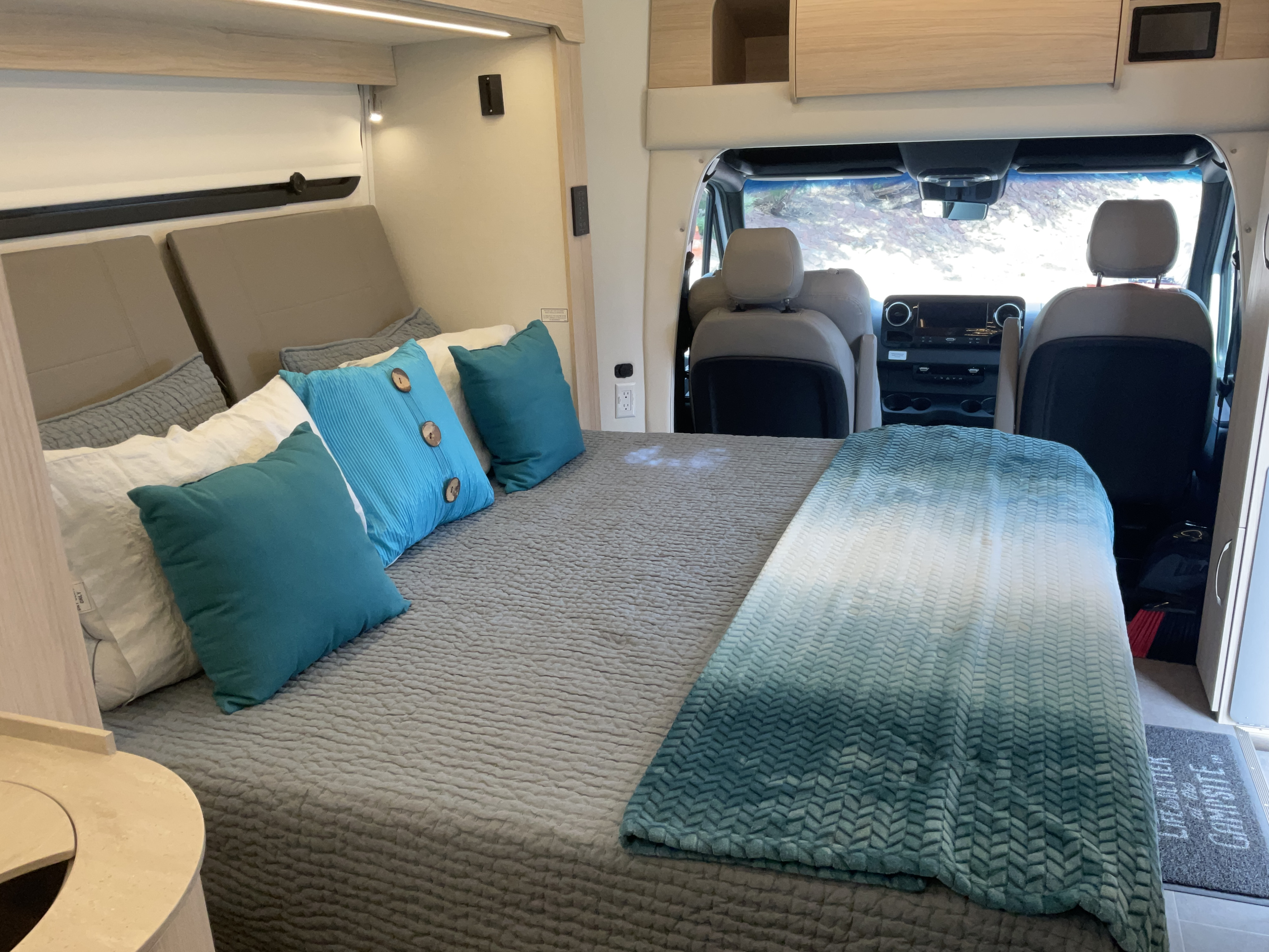 An RV rental's cozy sleeping area features a bed with teal accents and a view into the driver's cabin.. Leisure Travel Vans Unity 2024