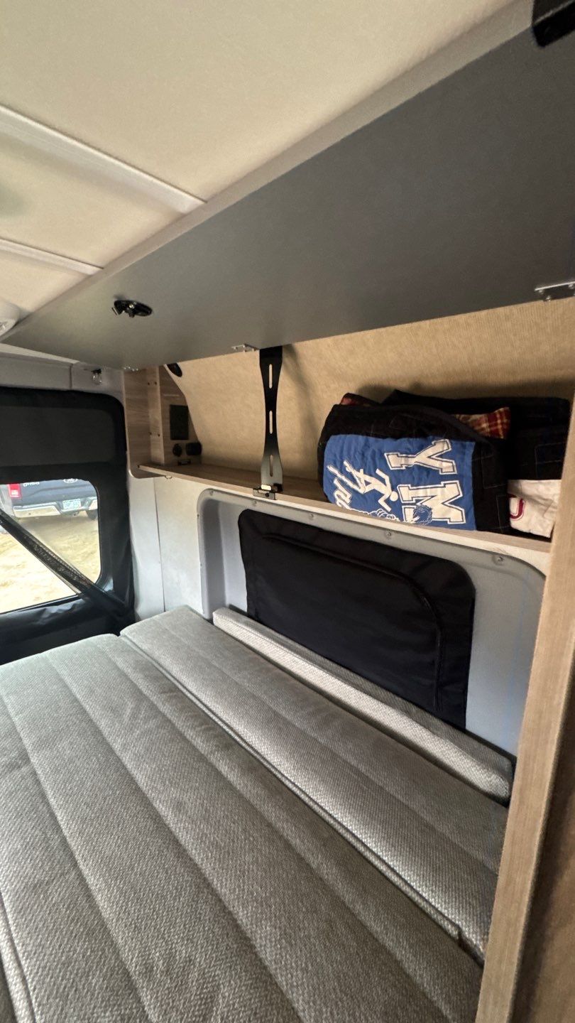 View inside an RV rental: a grey upholstered bed, window showing outdoors, and shelf storage with travel bags.. Winnebago Solis 2023