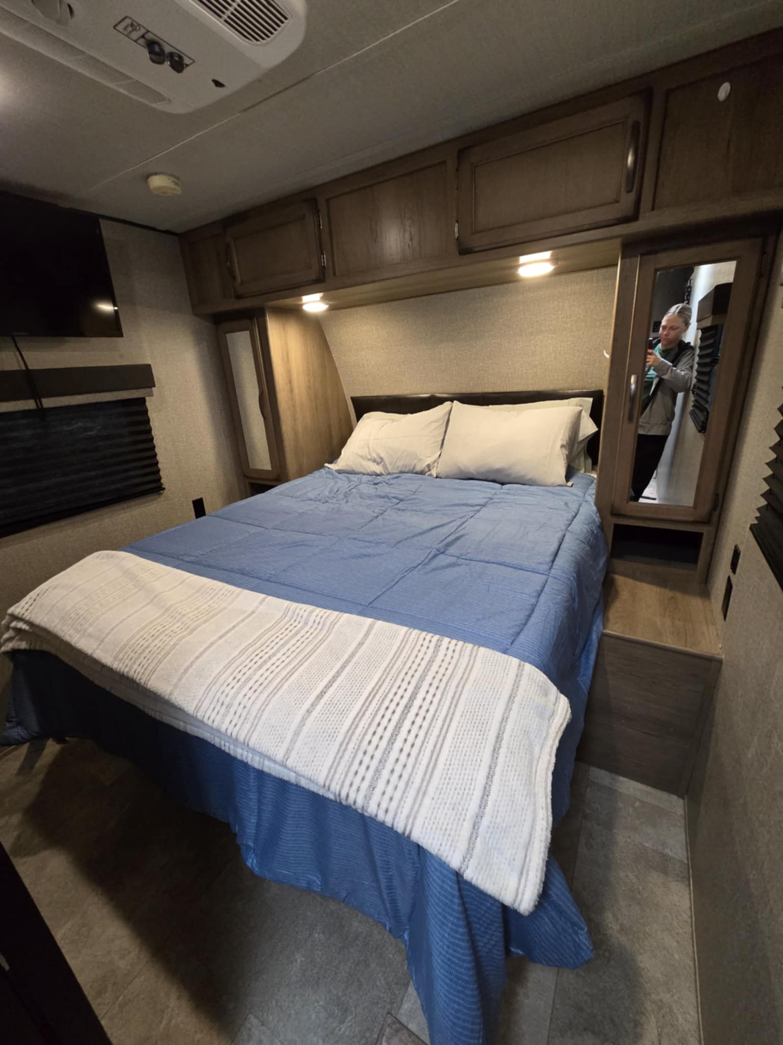 Cozy RV rental bedroom featuring a queen bed with blue bedding, overhead cabinets, and a mirror showing a reflection.. Grand Design Transcend 2019