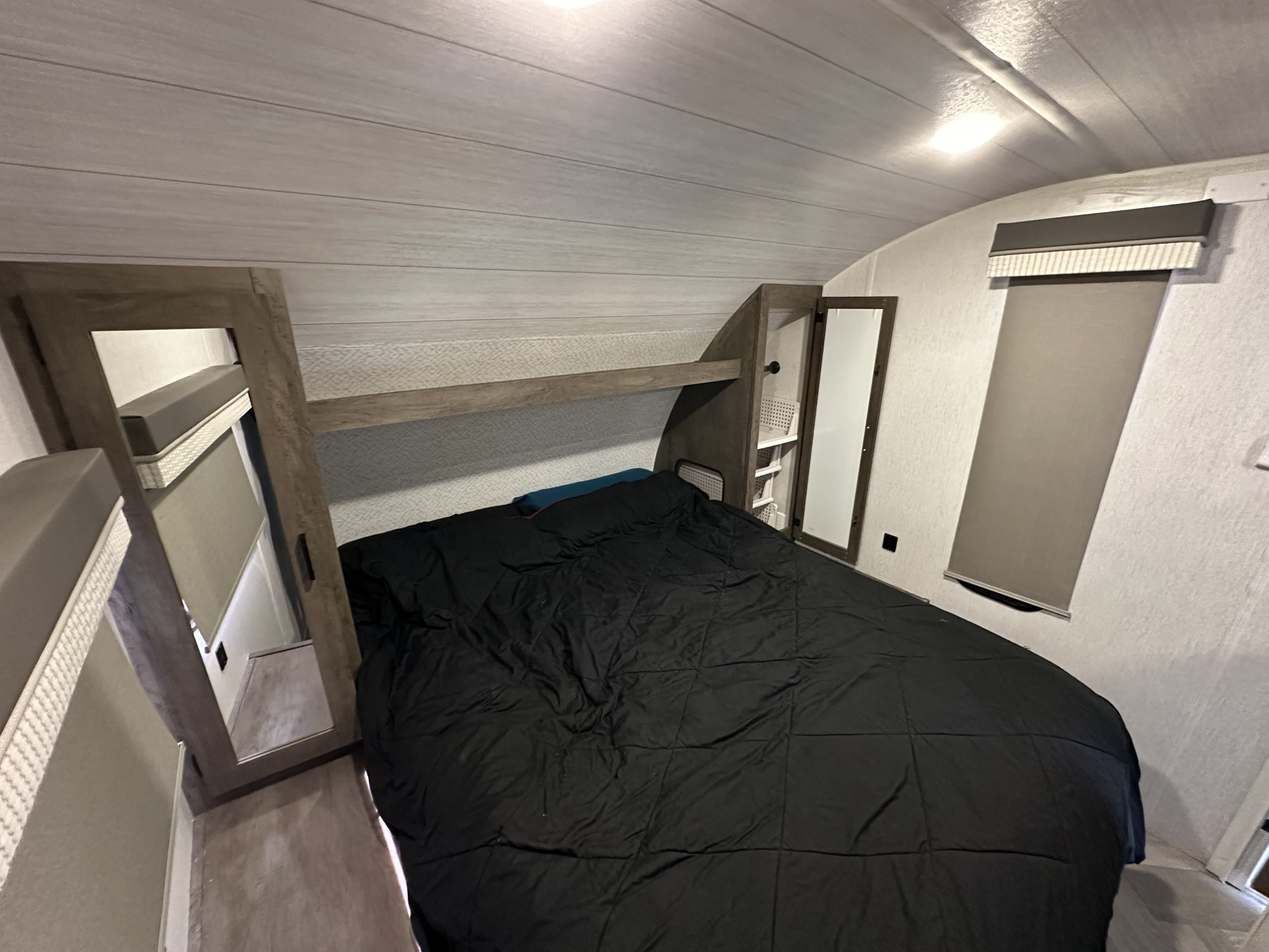 Cozy RV rental bedroom with a black bed, overhead storage, mirrored cabinet, and window blind.. Forest River Wildwood X-Lite 2022