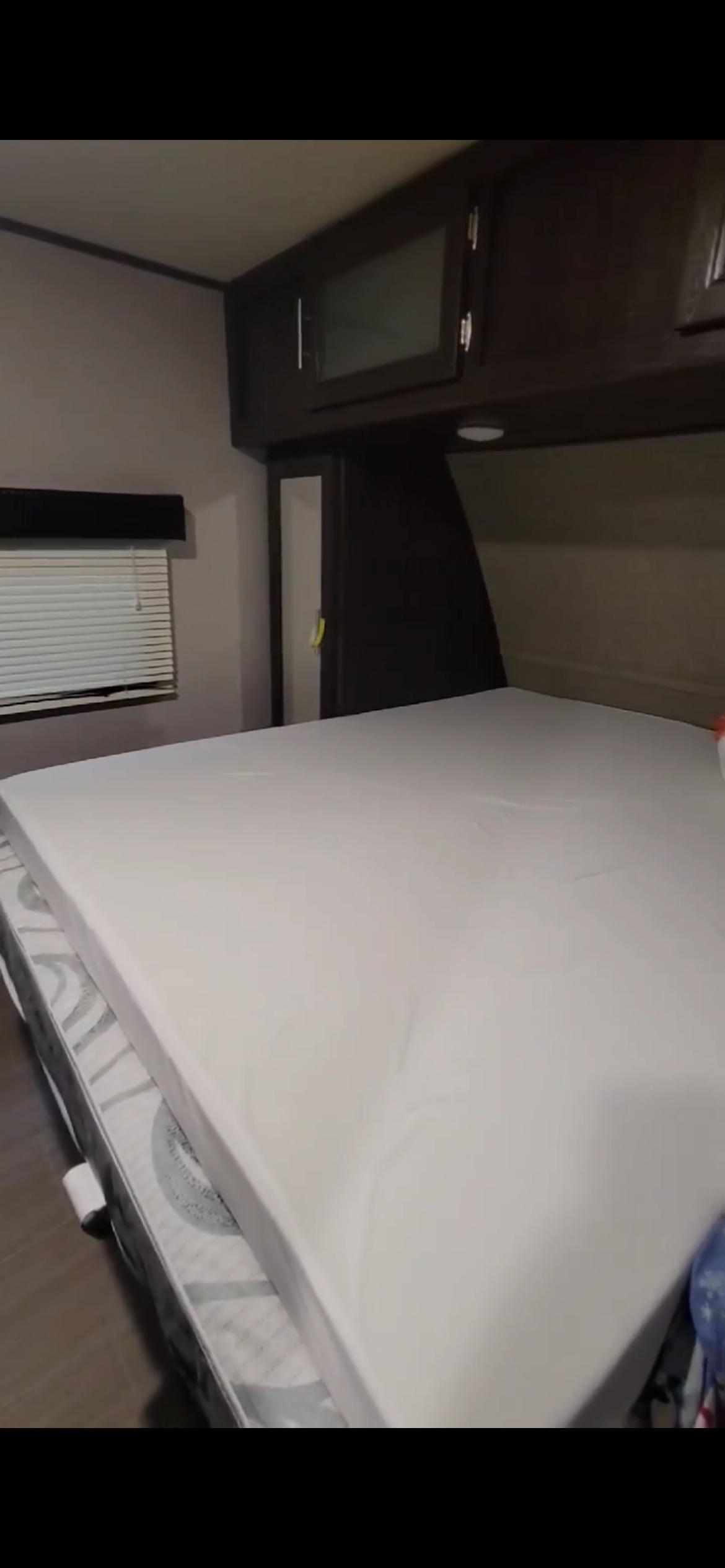 RV rental bedroom interior featuring a bed with a white mattress topper, dark overhead cabinets, and a window with blinds.. Dutchmen Kodiak Express 2018