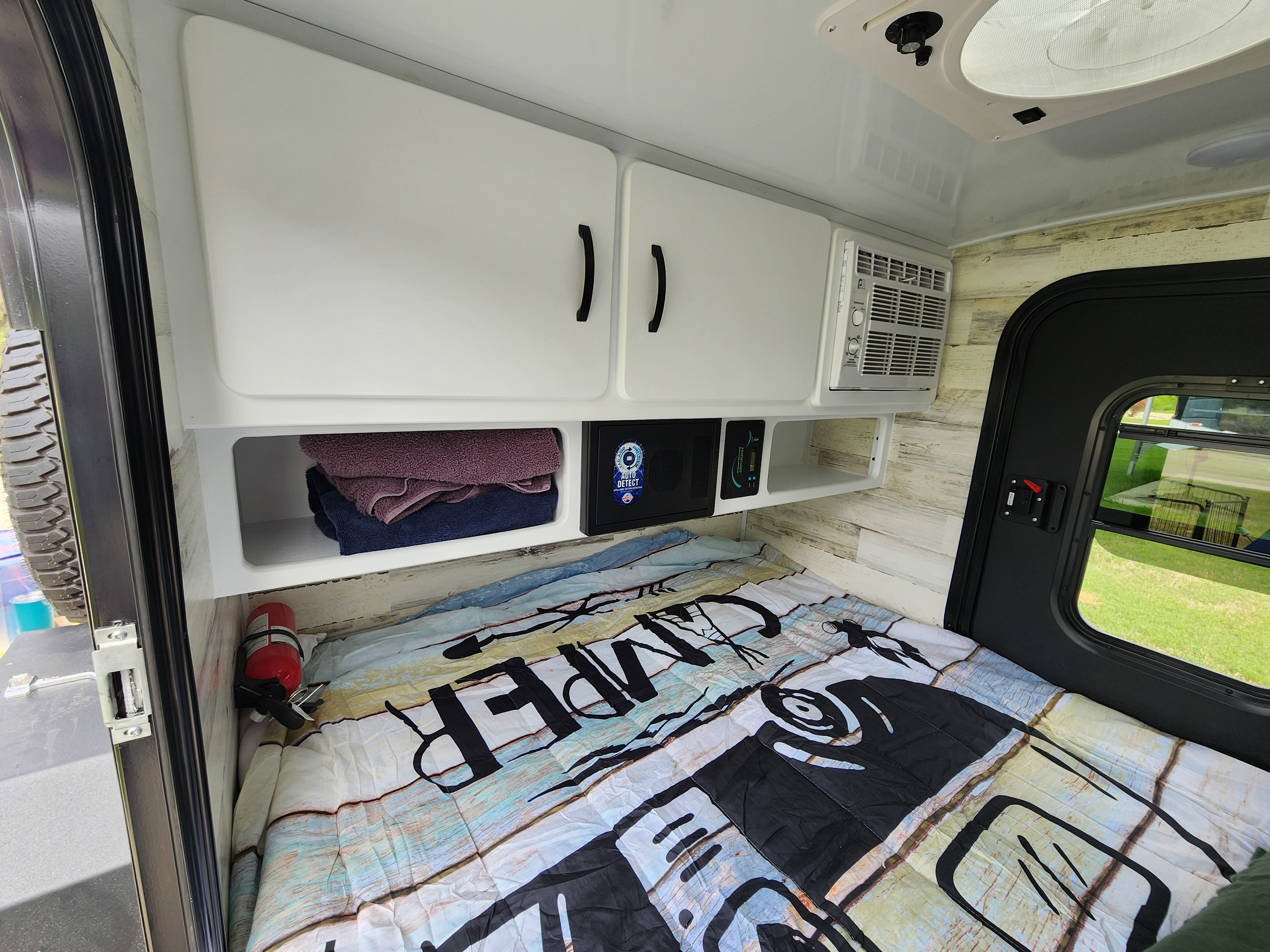 RV rental bedroom view: bed with 'CAMPER' comforter, wood walls, overhead cabinets, AC, and a security box.. Modern Buggy Lil Buggy 10RK 2025
