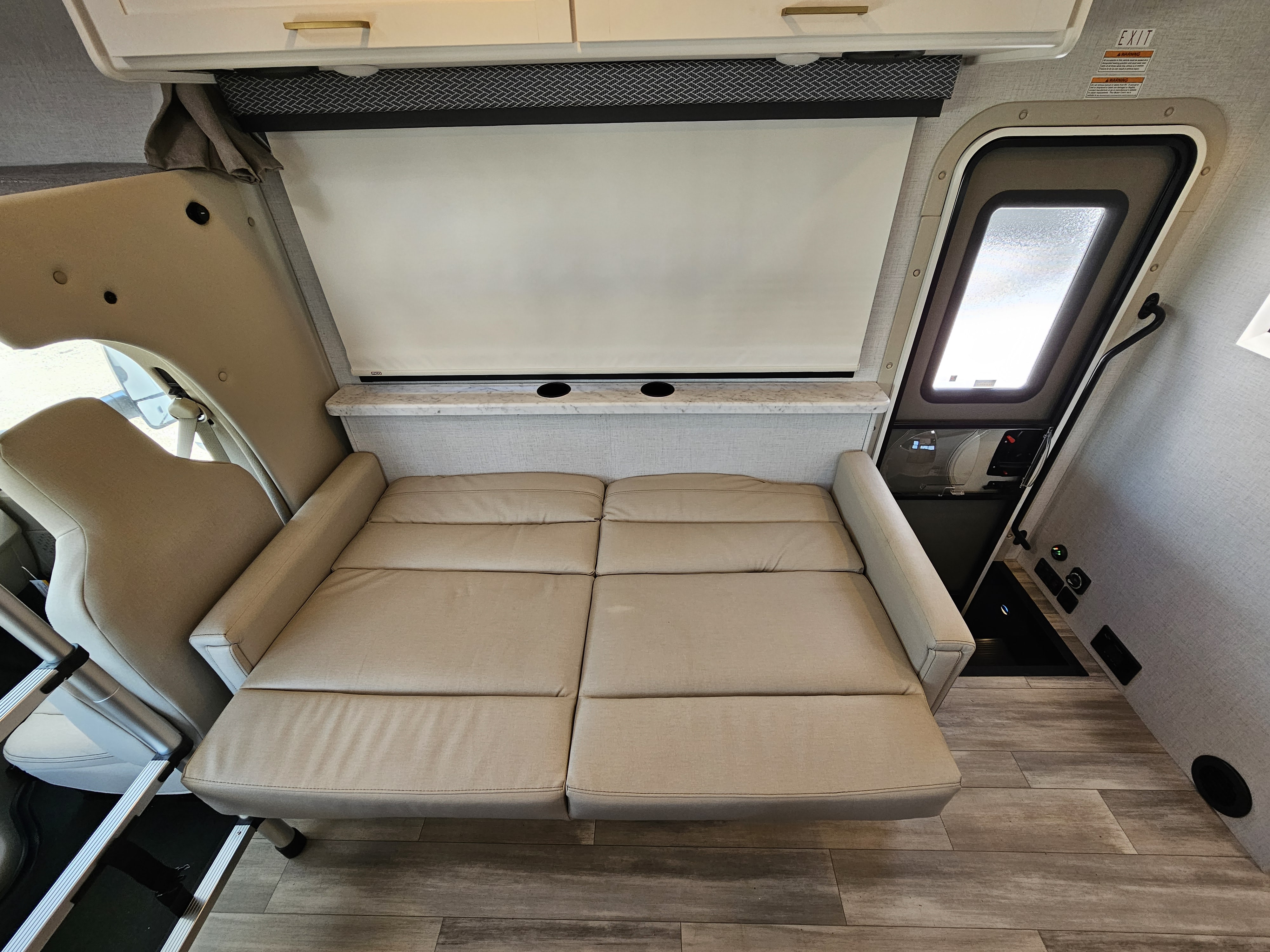 Inside an rv rental: versatile light-colored sofa converts to a bed, with cup holders, overhead storage, and an exit door.. Thor Motor Coach Four Winds 2026