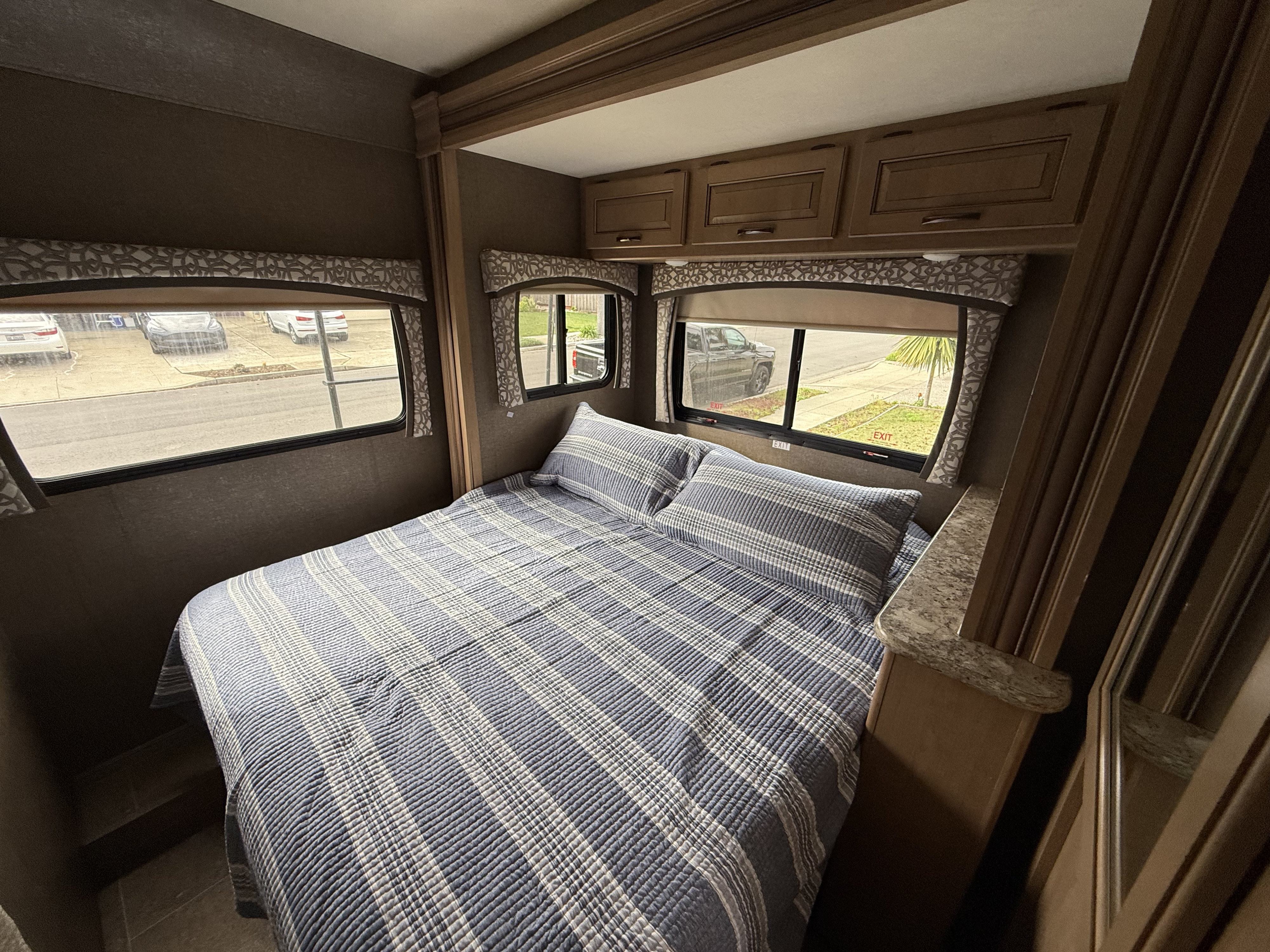 RV rental bedroom with a striped bed, multiple windows, and wooden overhead storage cabinets for travel.. Thor Chateau Sprinter 2018