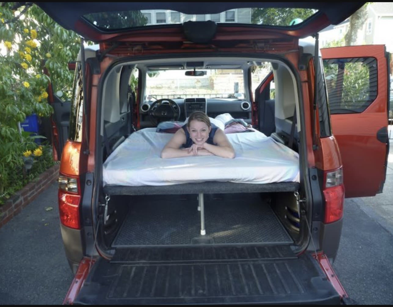 An orange Honda Element, modified as a DIY RV rental sleeper, features a cozy bed with a smiling woman inside.. Honda Element 2004